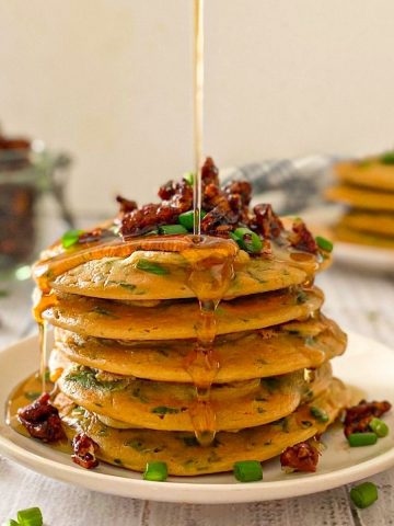 Stack of pancakes with bacon and chives on top and syrup dripping down.