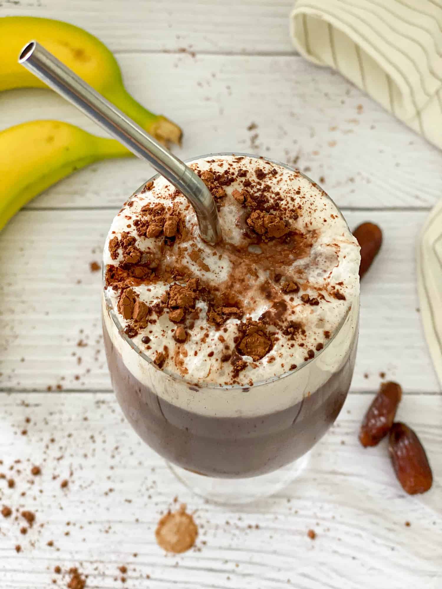 Metal straw inside glass of chocolate beverage with dates and bananas around it.
