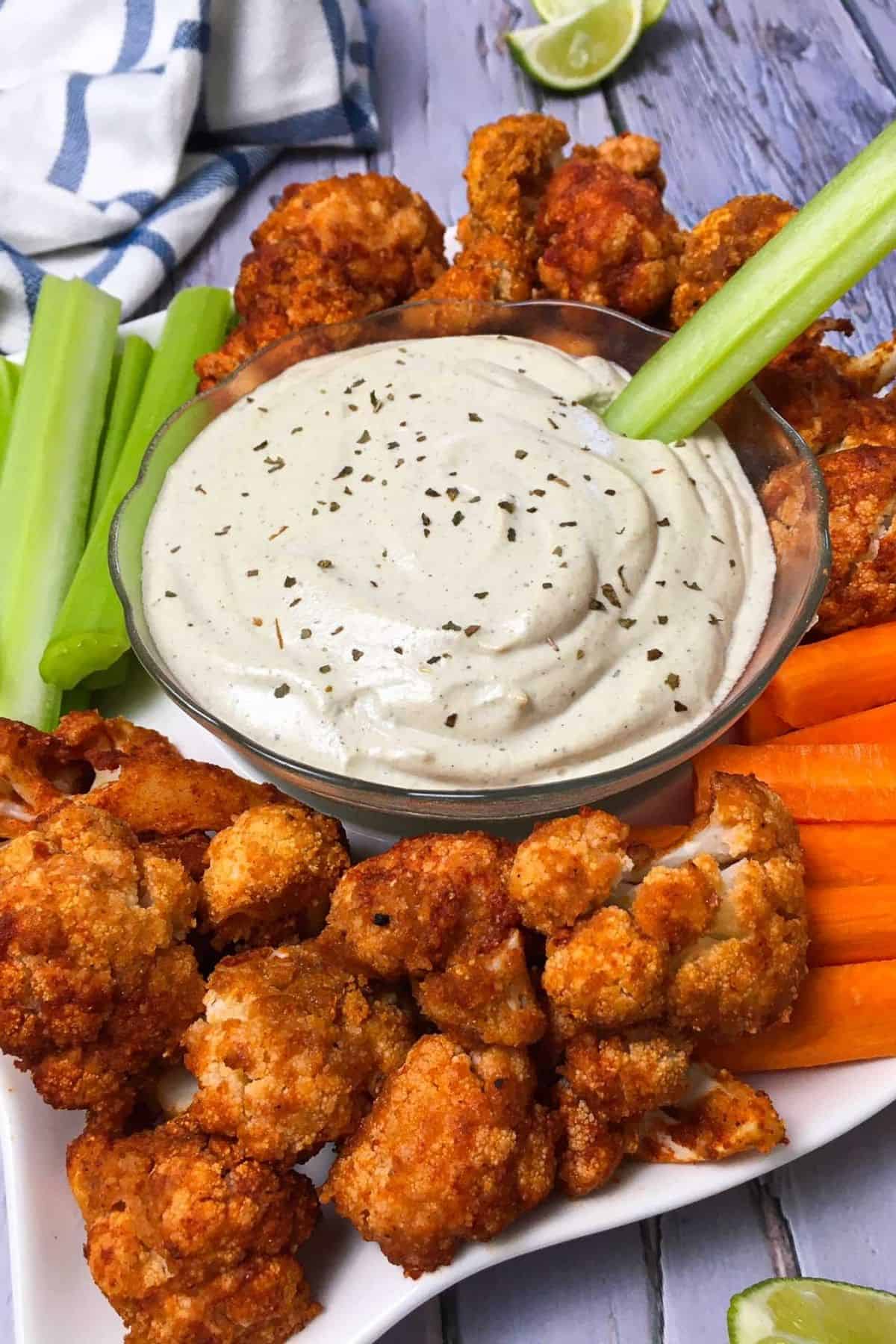 White platter with buffalo cauliflower, carrots and celery sticks with white dip in the center.