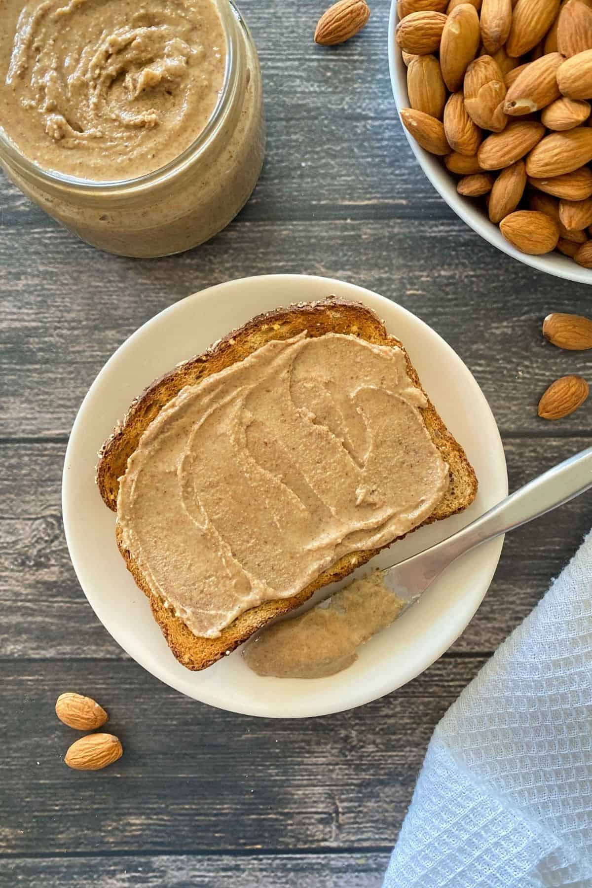Toast with almond butter and knife beside it on white plate with bowl of almonds in background.