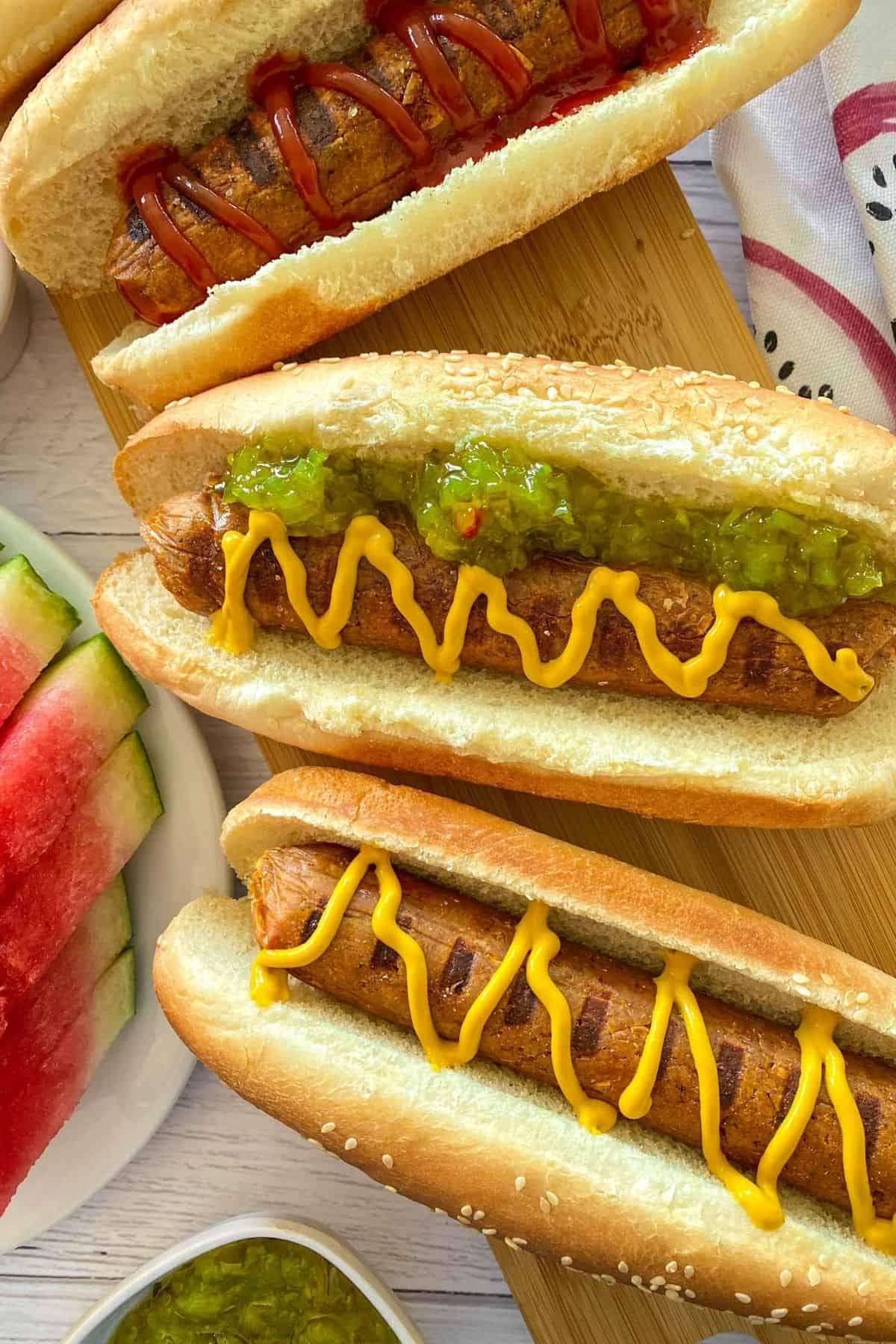 Seitan hot dogs in buns with mustard, relish and ketchup on cutting board.