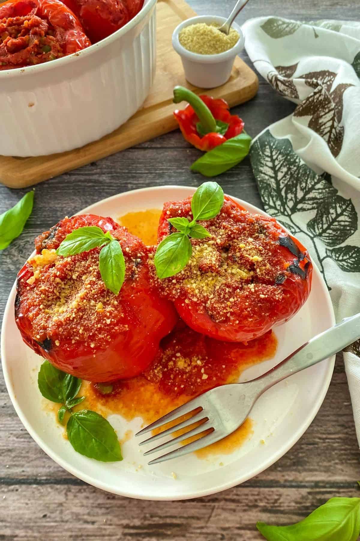 Two whole stuffed red peppers on white plate with fork on the side and basil on top of peppers.