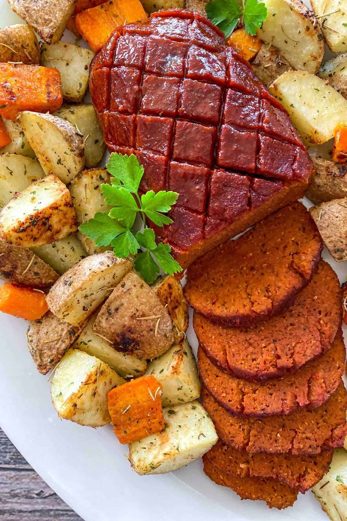 A vegan ham sliced on platter with roasted potatoes and carrots surrounding it.