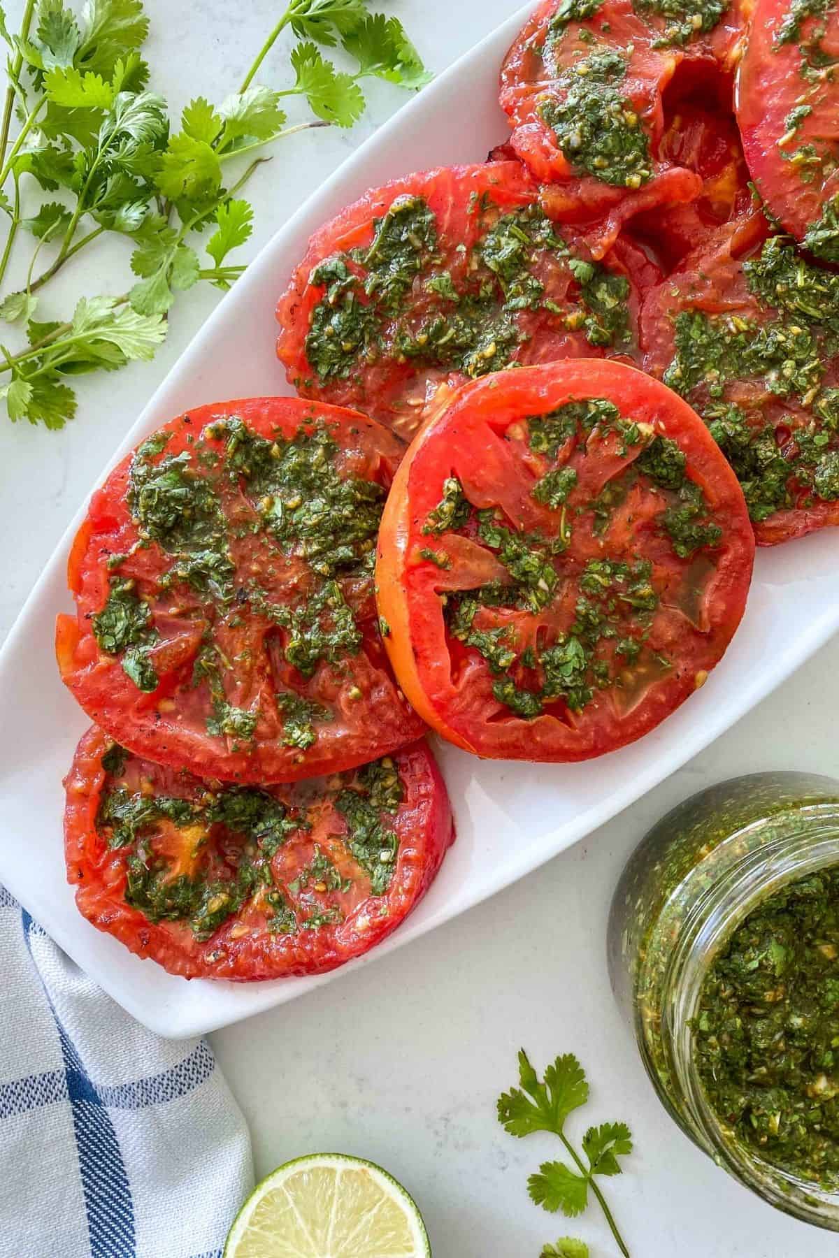 White platter filled with sliced grilled tomatoes with chimichurri on top.