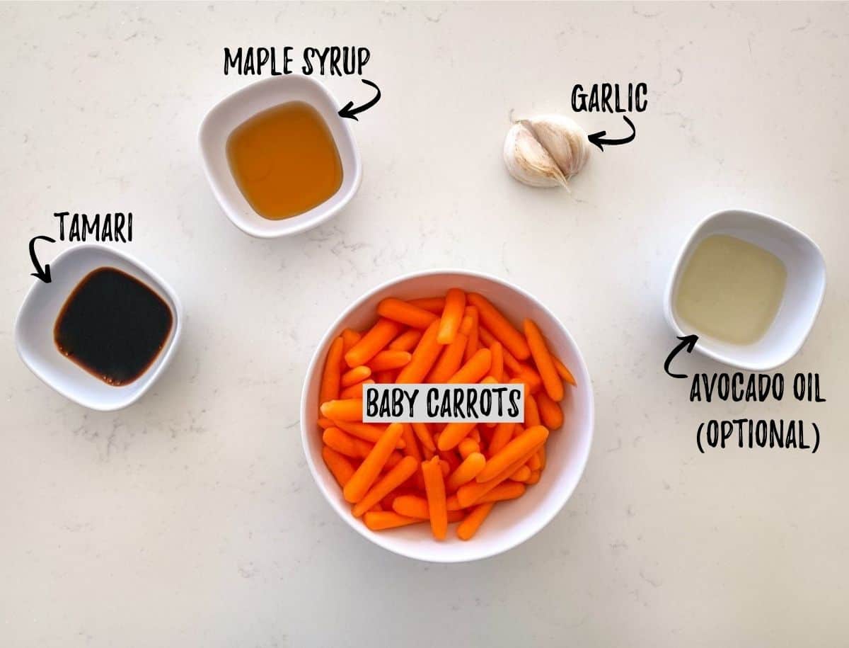 Baby carrots, tamari, maple syrup and oil in small prep bowls on counter with garlic cloves beside.