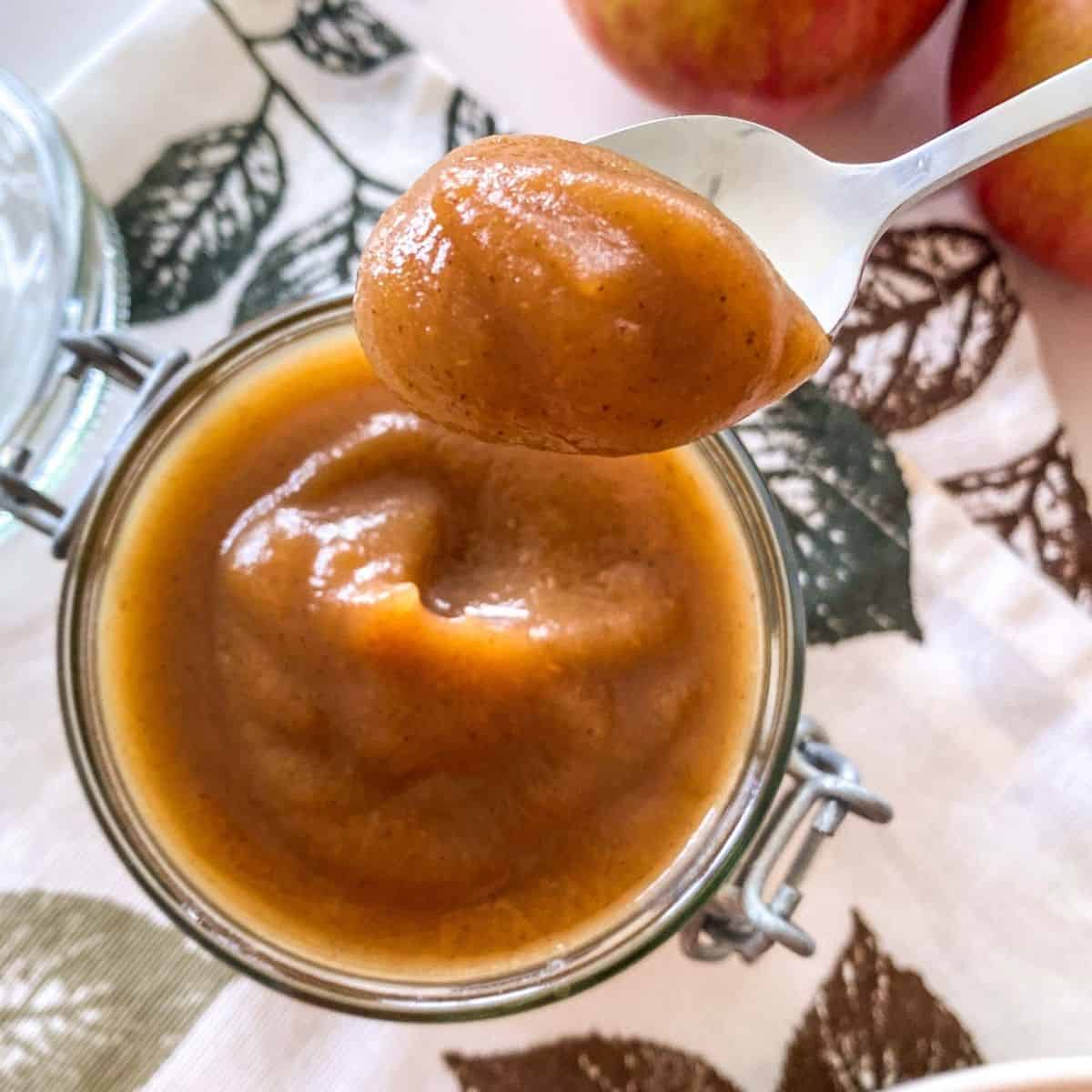 Jar of apple butter with spoonful held up above it and apples in the background.