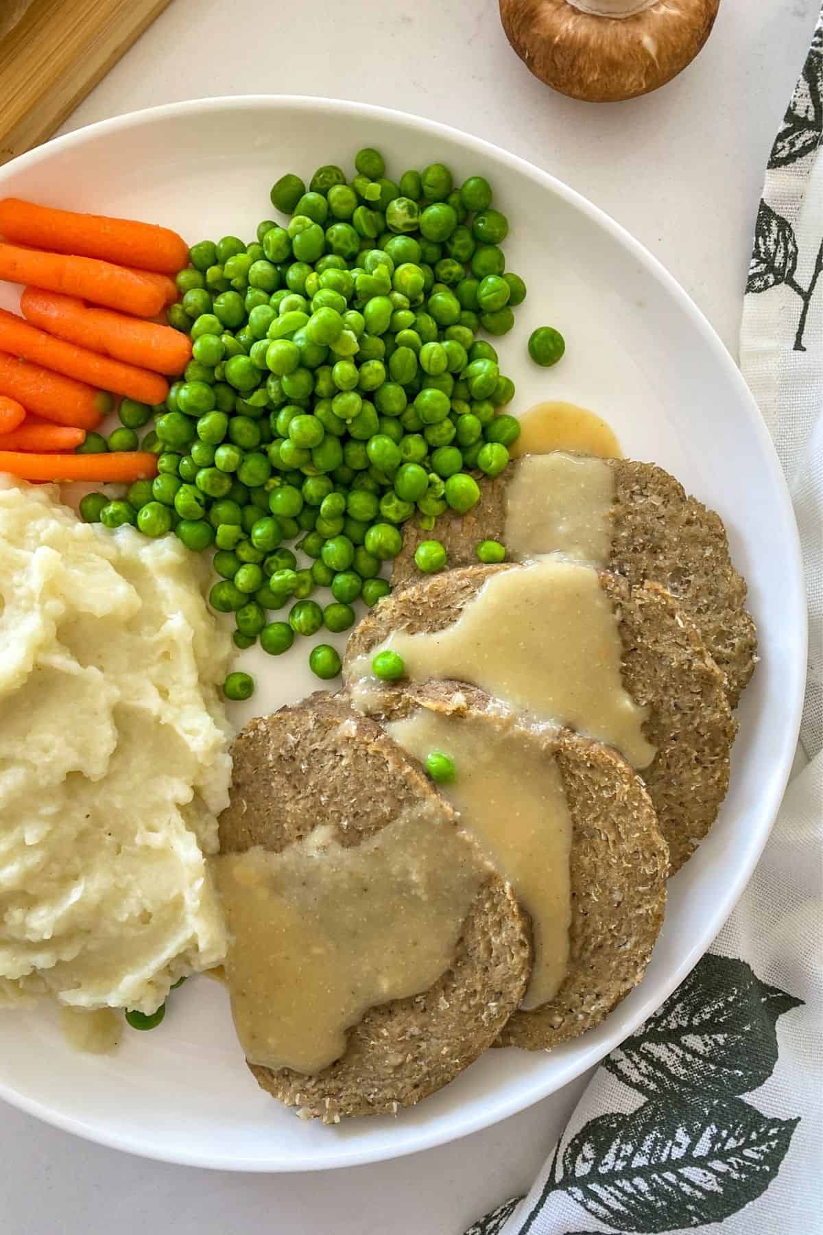 White plate with slices of meat, peas, carrots and mashed potaoes.