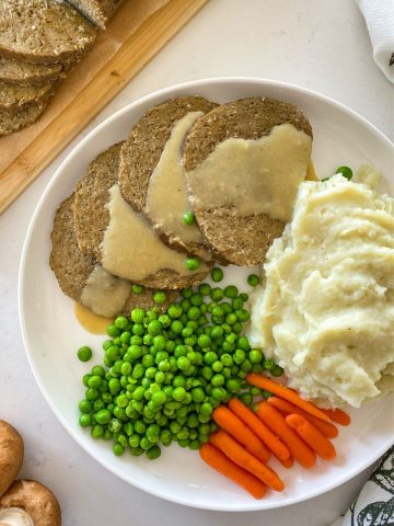 Sliced roast with gravy on top and peas, carrots and mashed potatoes beside it.