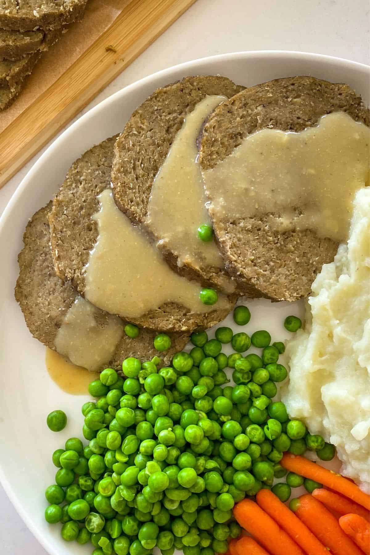 Vegan turkey roast slices on white plate with mashed potatoes, peas and carrots.