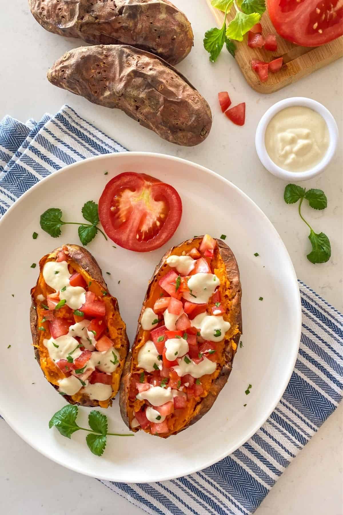 Baked sweet potatoes in white plate with tomato and sour cream and whole cooked sweet potatoes in background.
