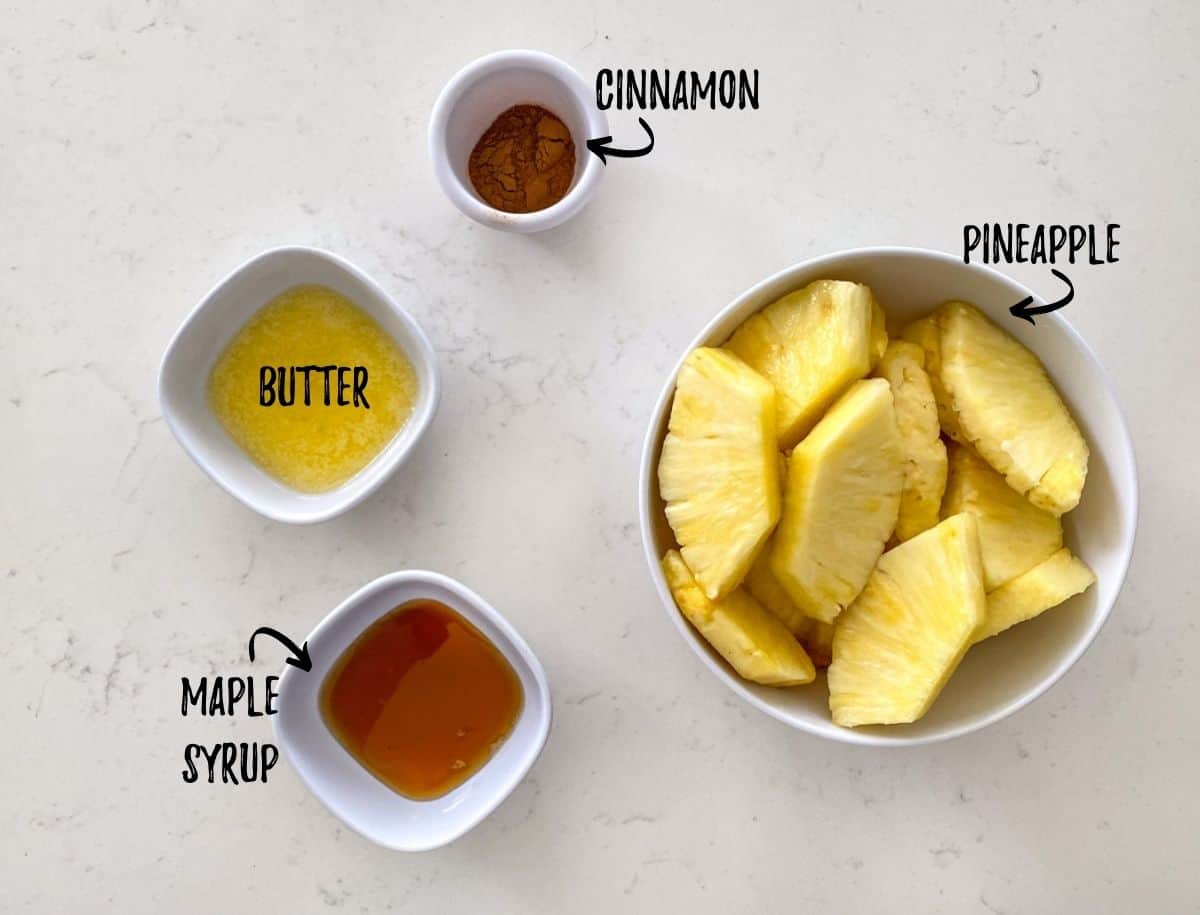 Bowl of pineapple slices, bowl of maple syrup, butter and cinnamon on counter top.