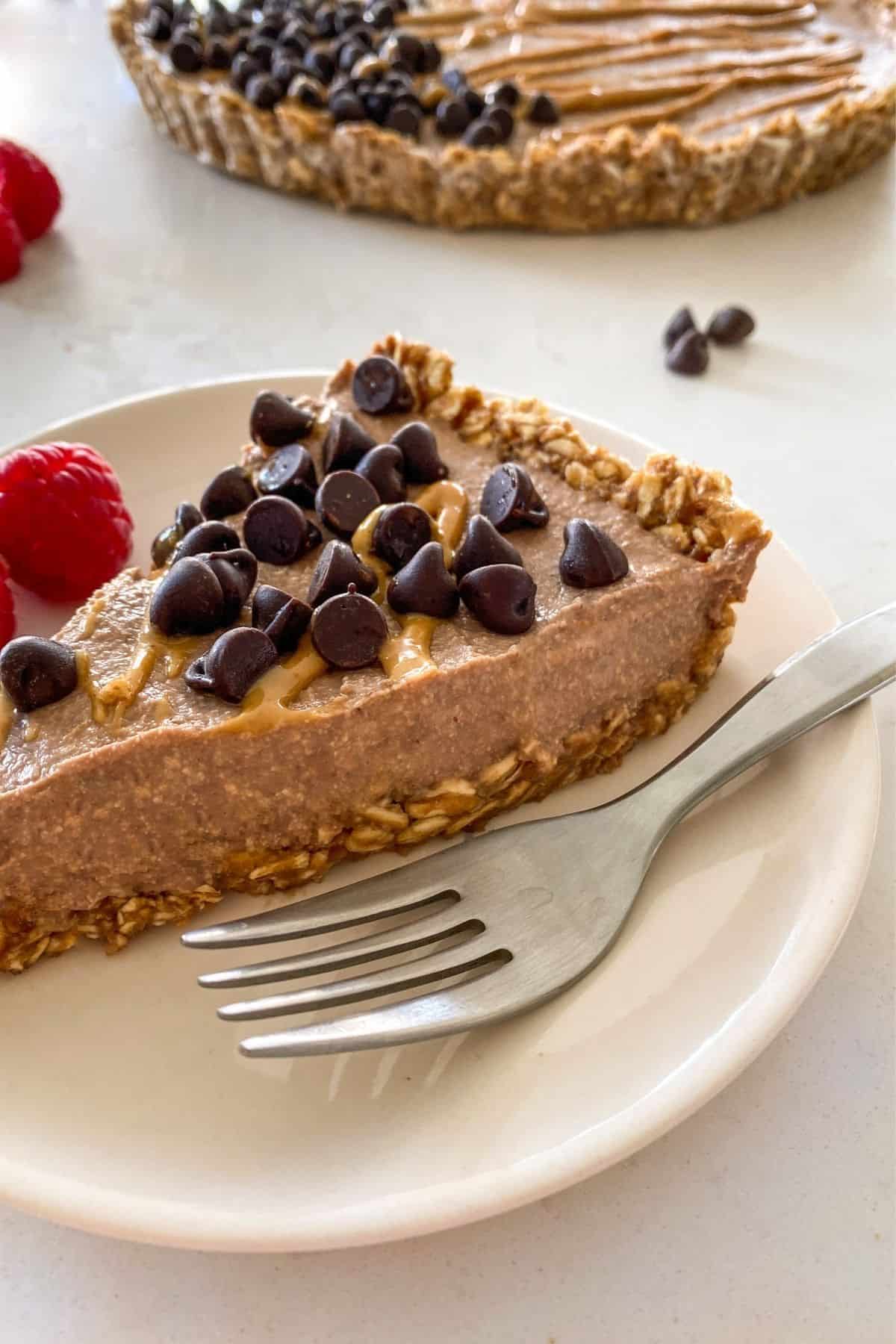 Slice of peanut butter pie in plate with fork and chocolate chips on top with raspberries on the side.