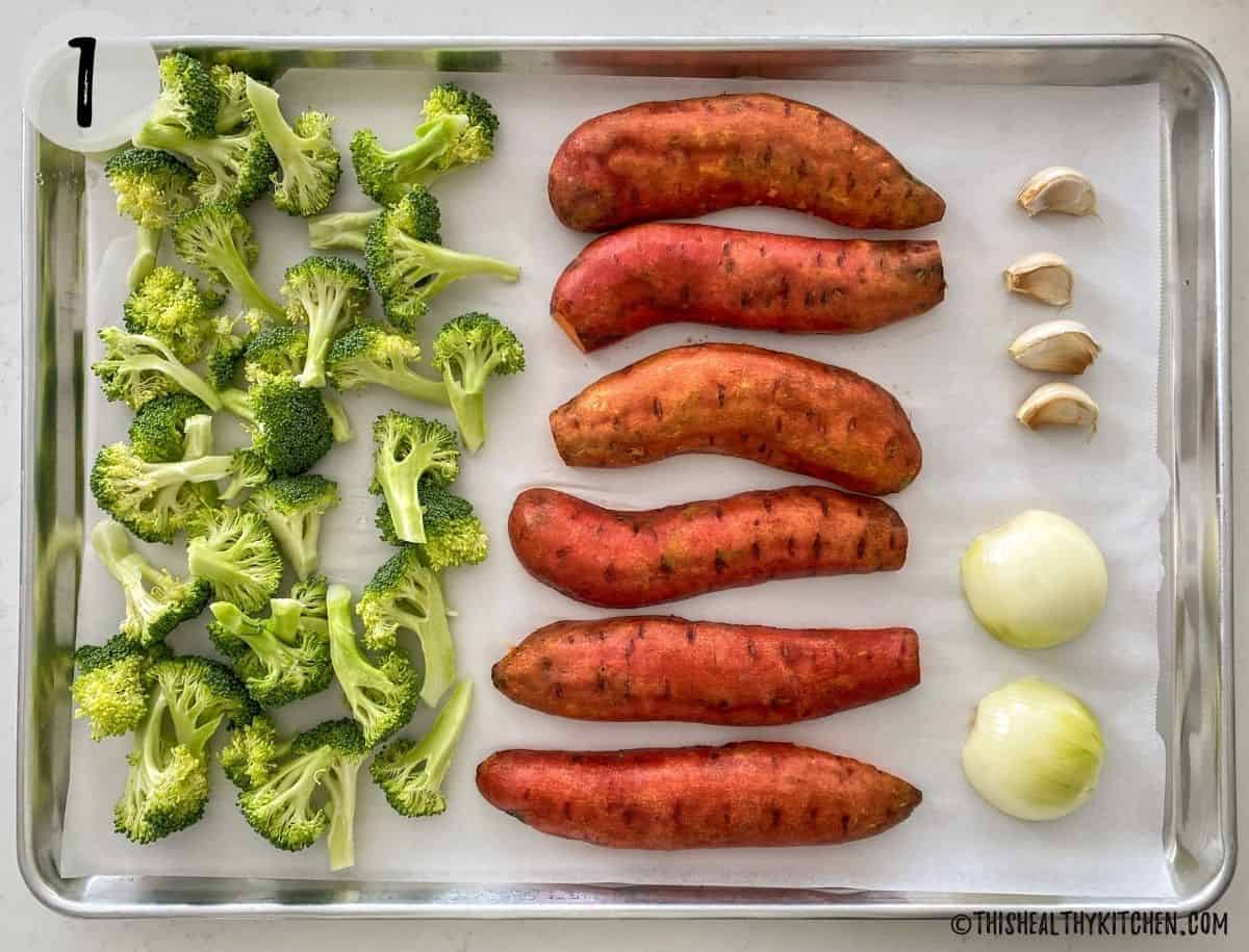 Large tray with broccoli florets, halved sweet potatoes, halved onion and garlic cloves.