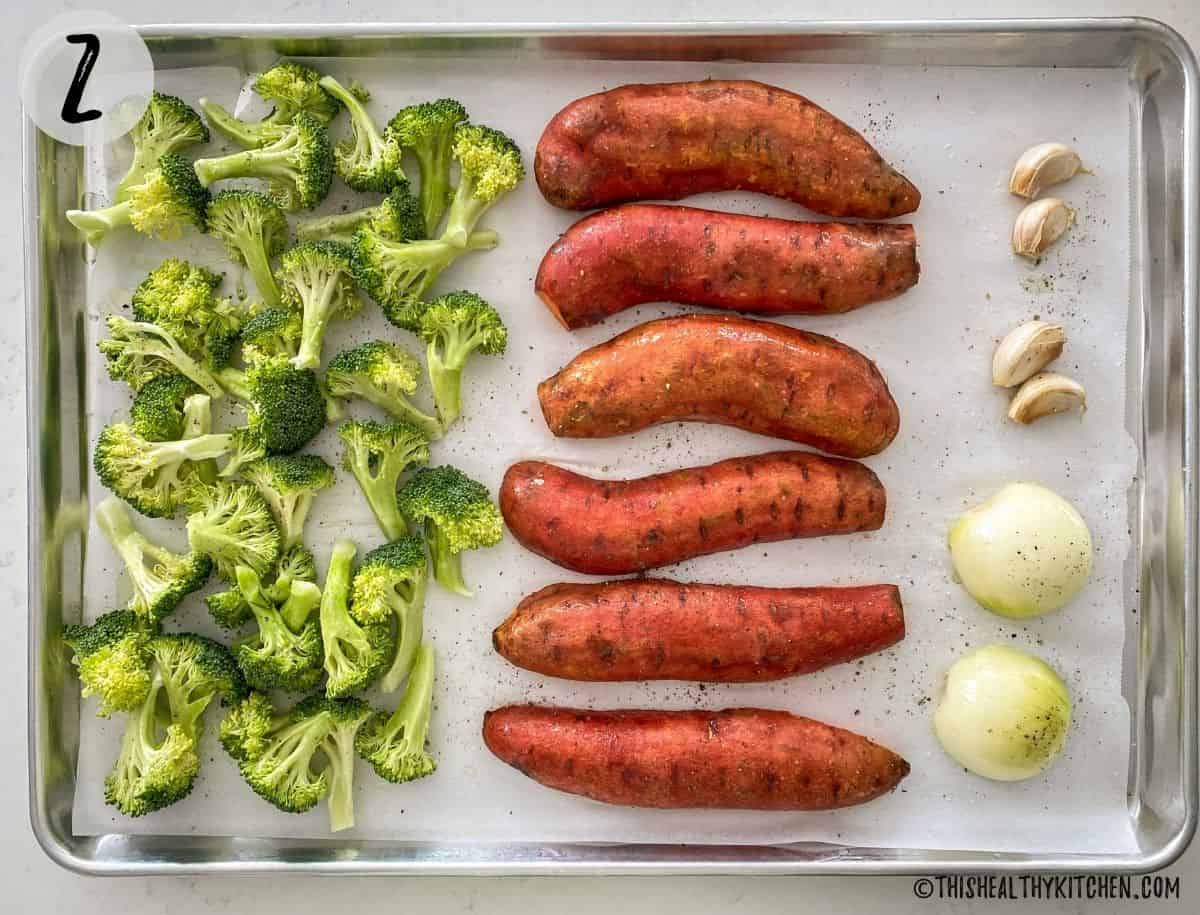 Large baking sheet with broccoli florets, halved sweet potatoes, halved onion and garlic cloves.