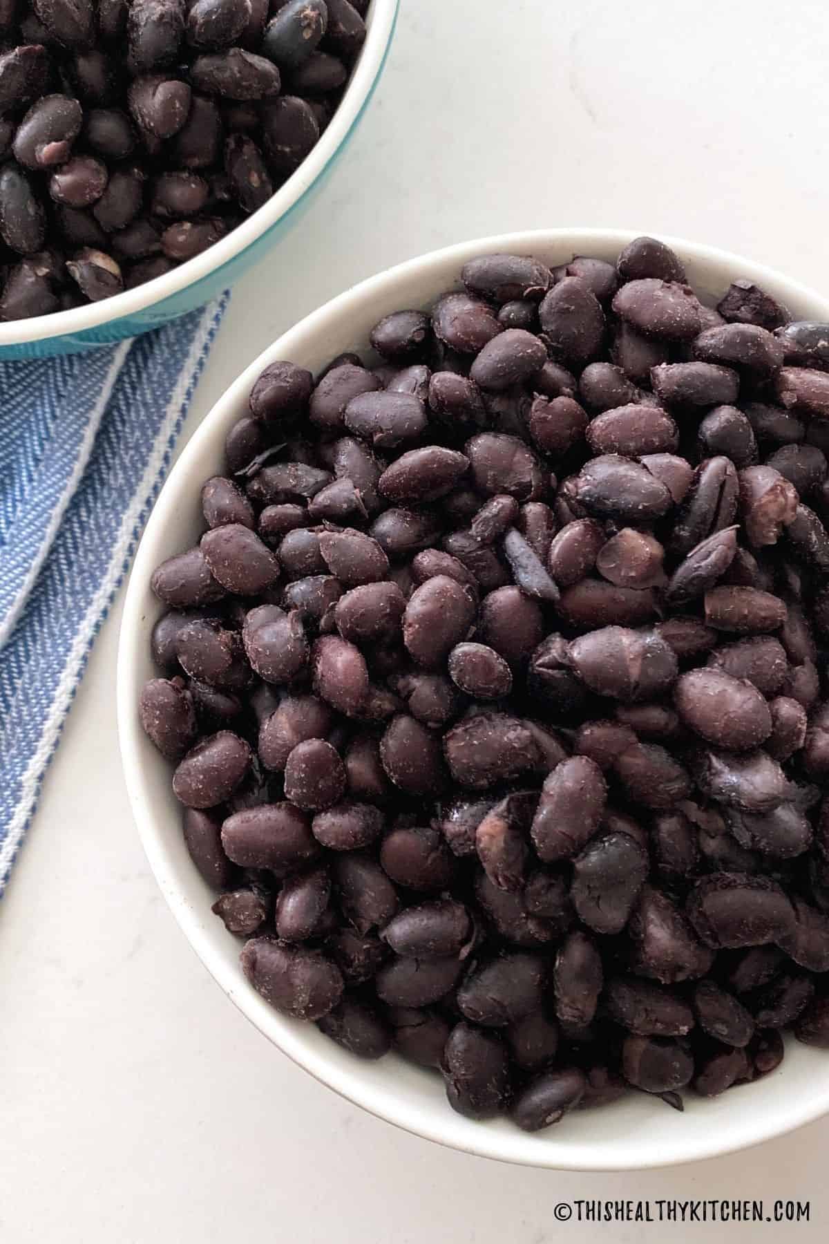 Two bowls of cooked black beans with blue dish towel on the side.
