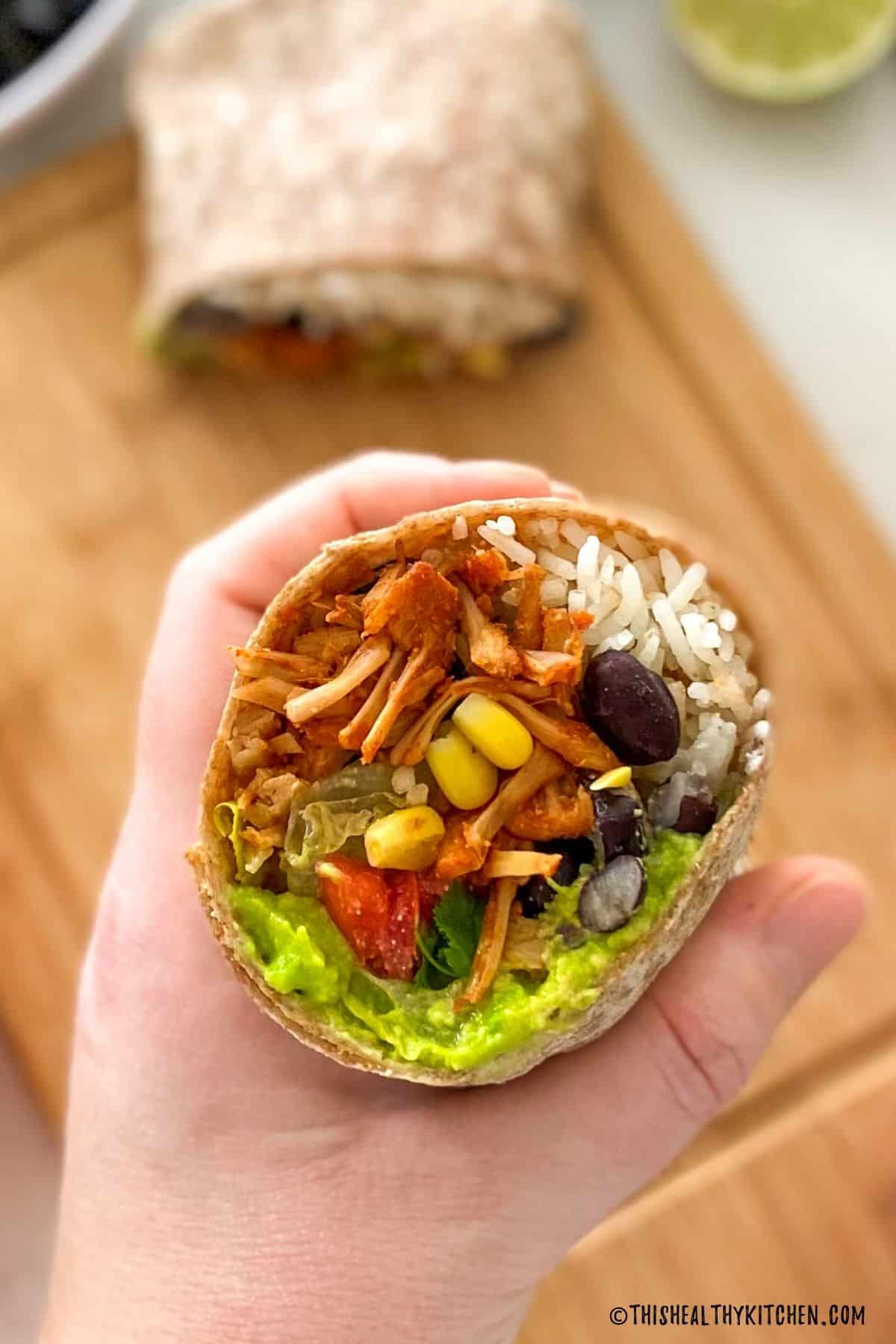 Hand holding up a vegan burrito that's been cut in half with remaining half on cutting board below.