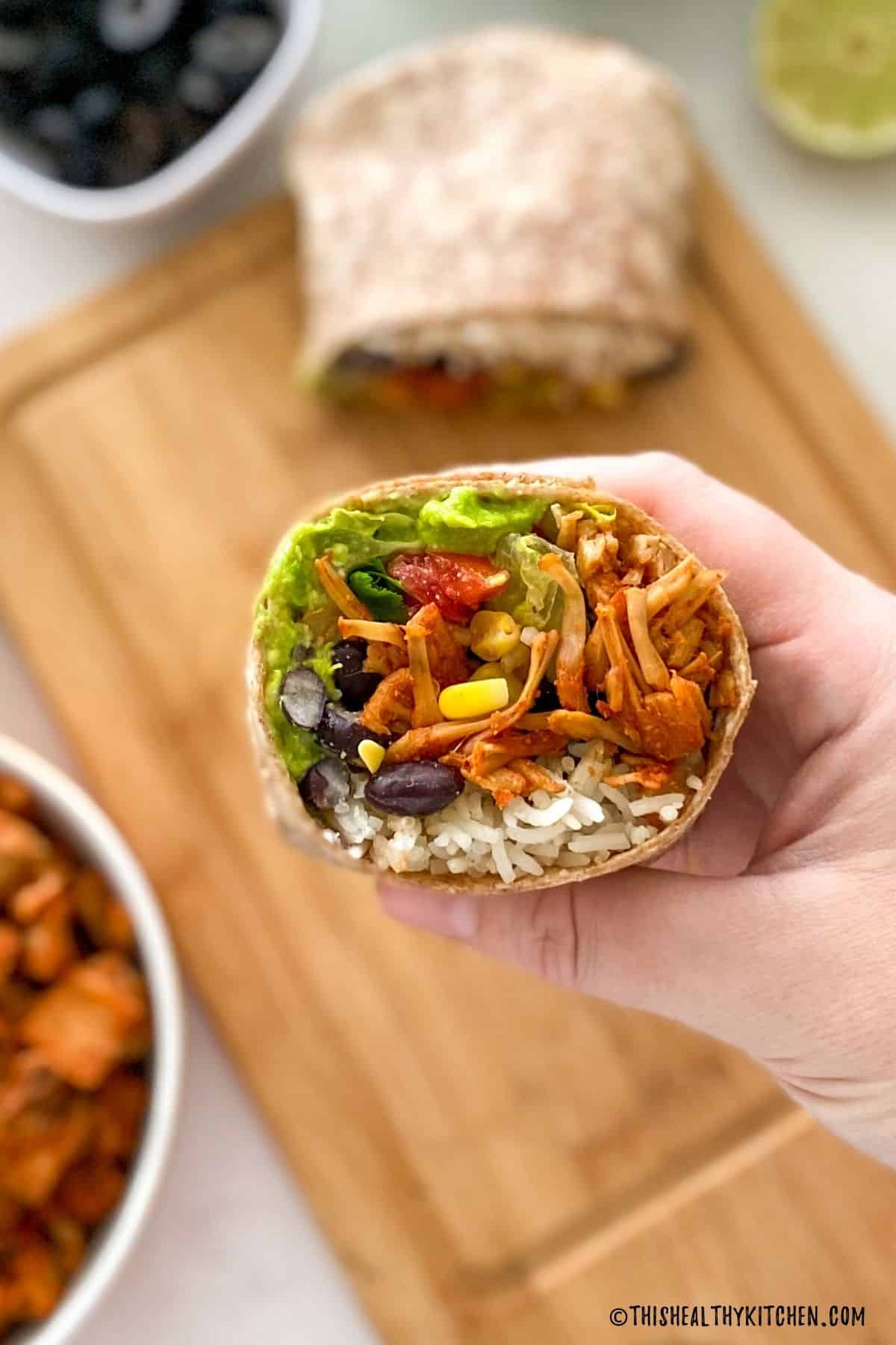 Halved vegan burrito held up in hand with remaining half on cutting board below.