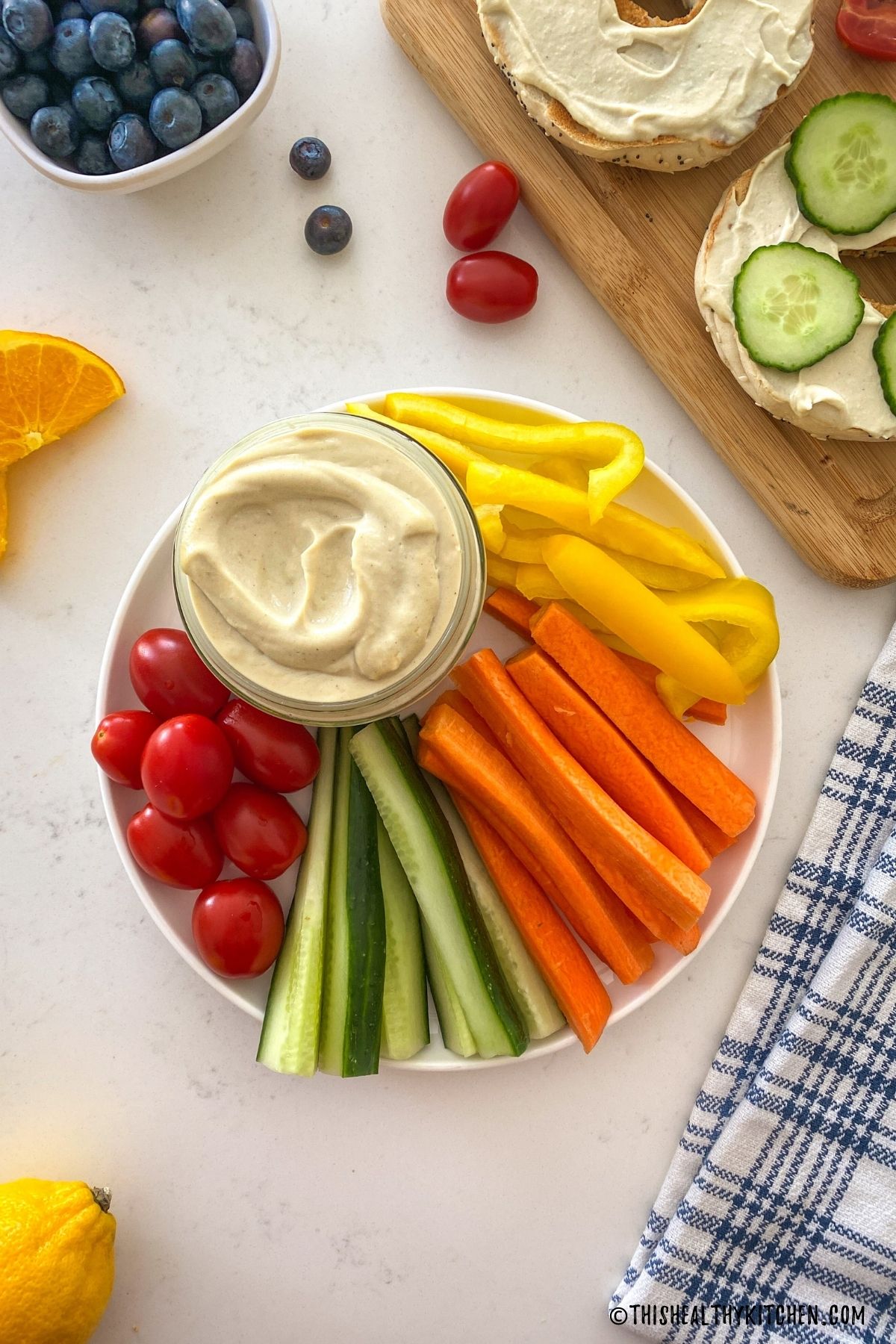 Jar of tofu cream cheese on plate with yellow peppers, carrot sticks, cucumber sticks and cherry tomatoes.
