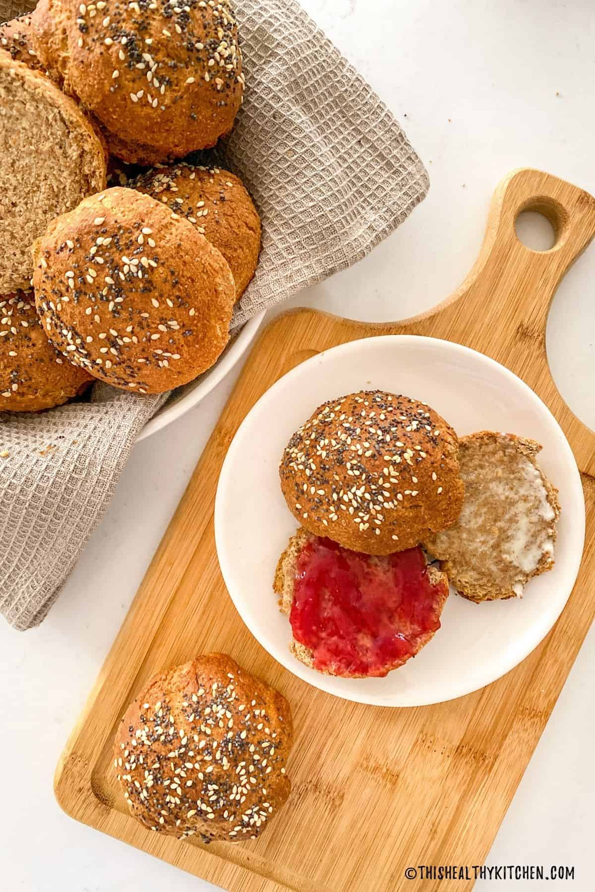 Hamburger buns in bread basket with one sliced open bun on plate with jam on one half and butter on the other.