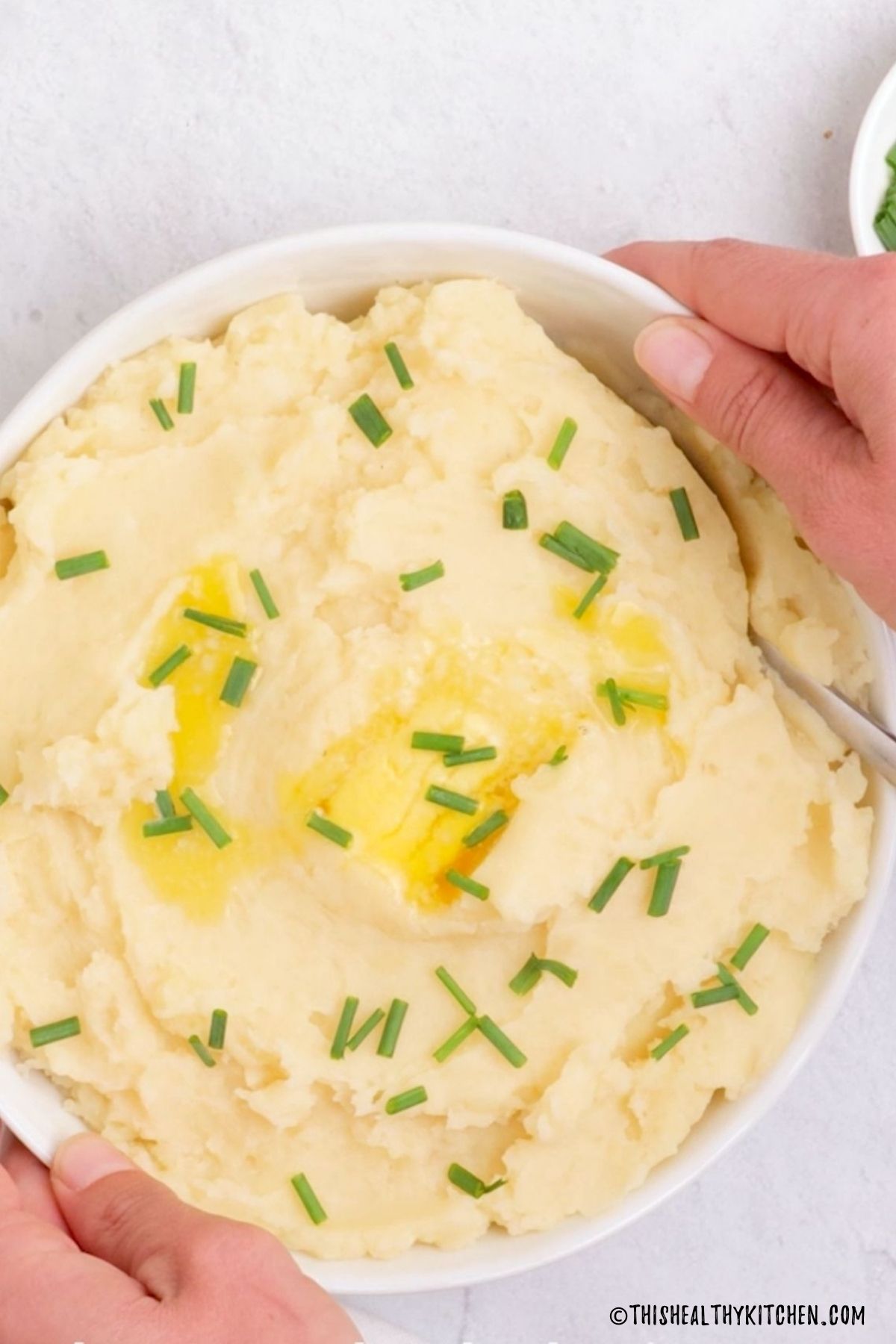 Hand holding up dish filled with mashed potatoes and garnished with chives.