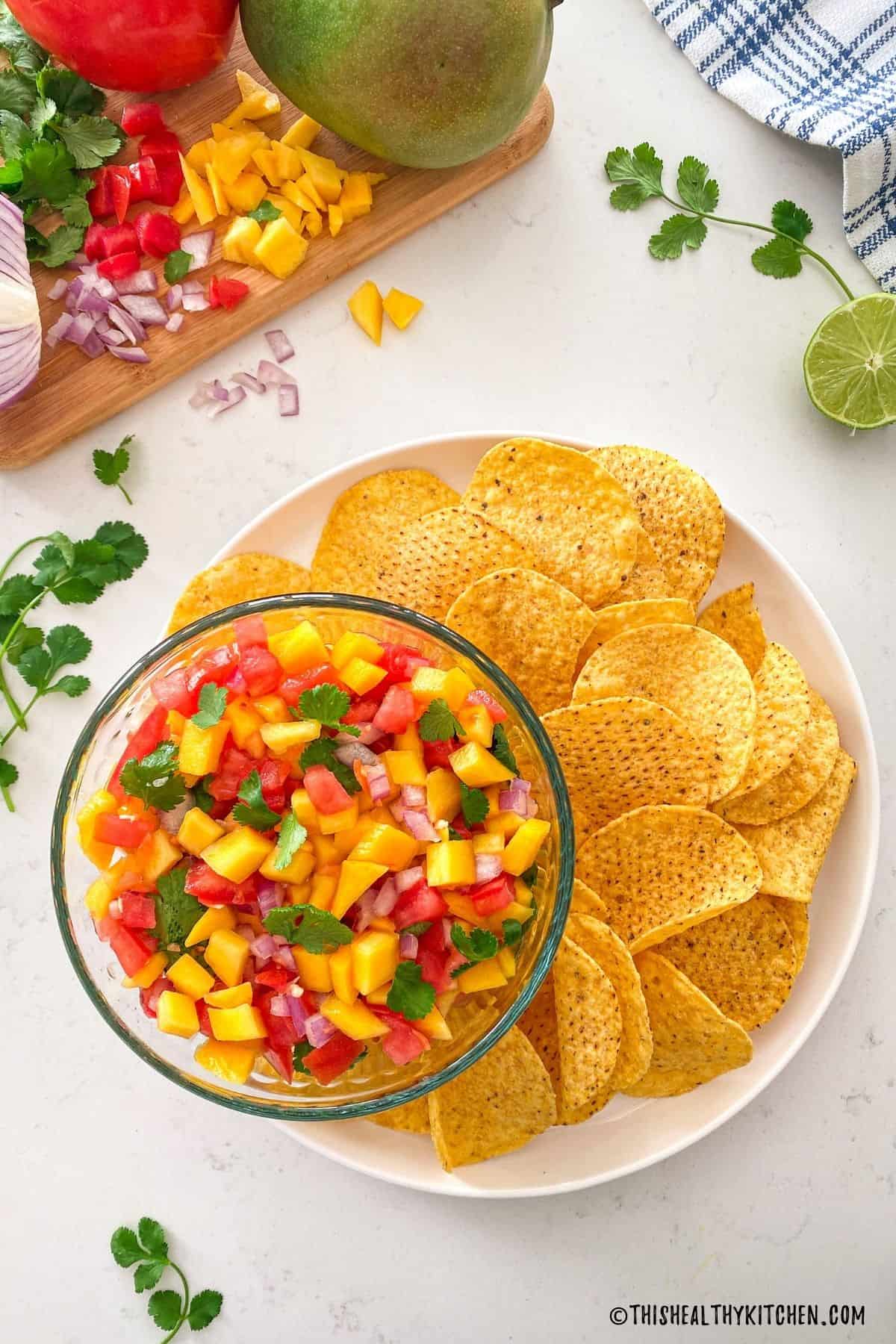 Bowl of mango salsa on platter with corn chips all around it.