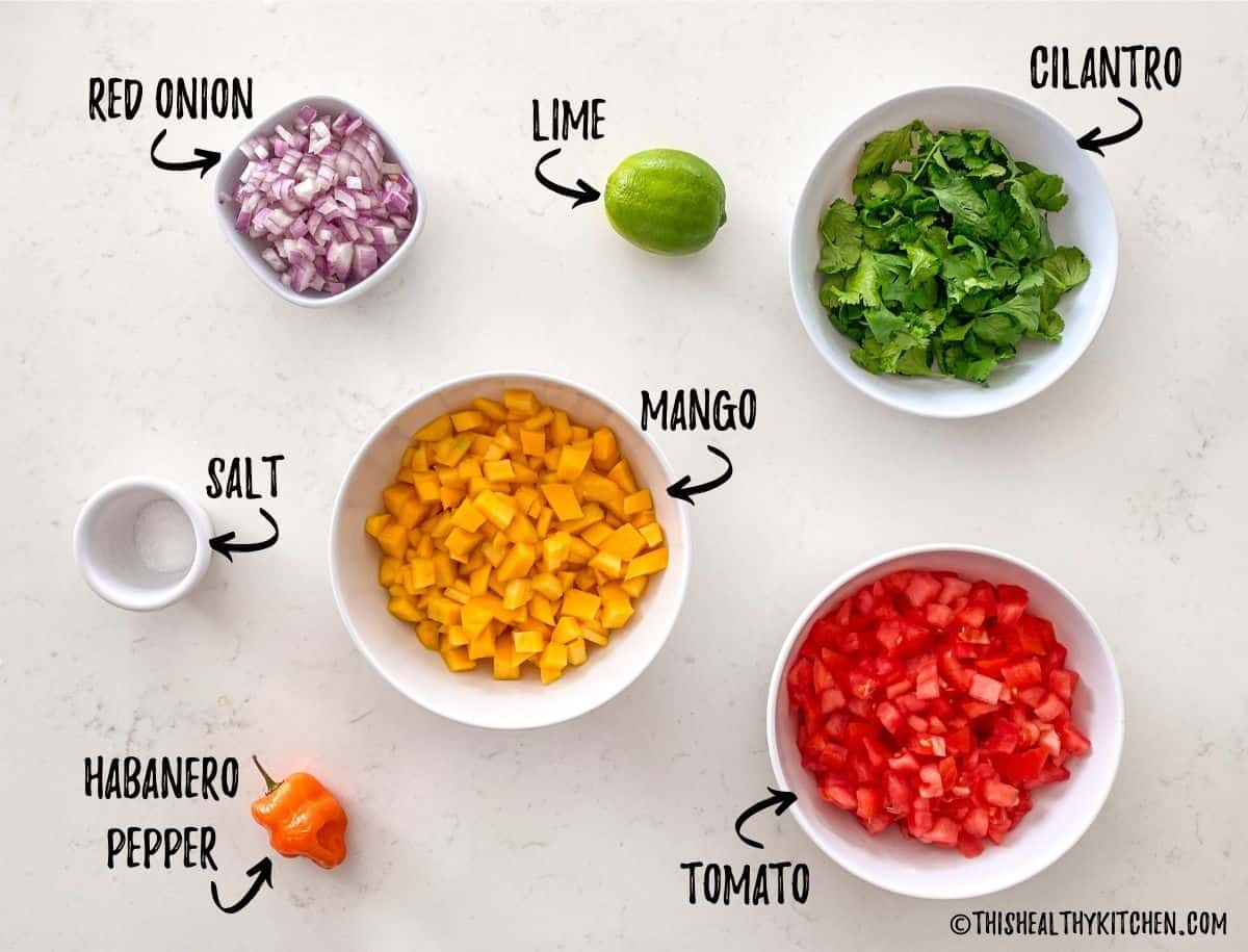 Tomato, mango, cilantro, onion, salt, lime and pepper on white kitchen counter.