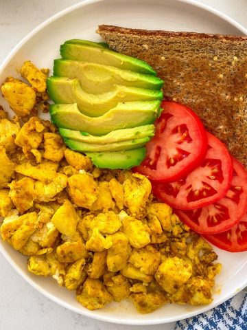 White plate with tofu scramble, avocado slices, tomato slices and a slice of toast.