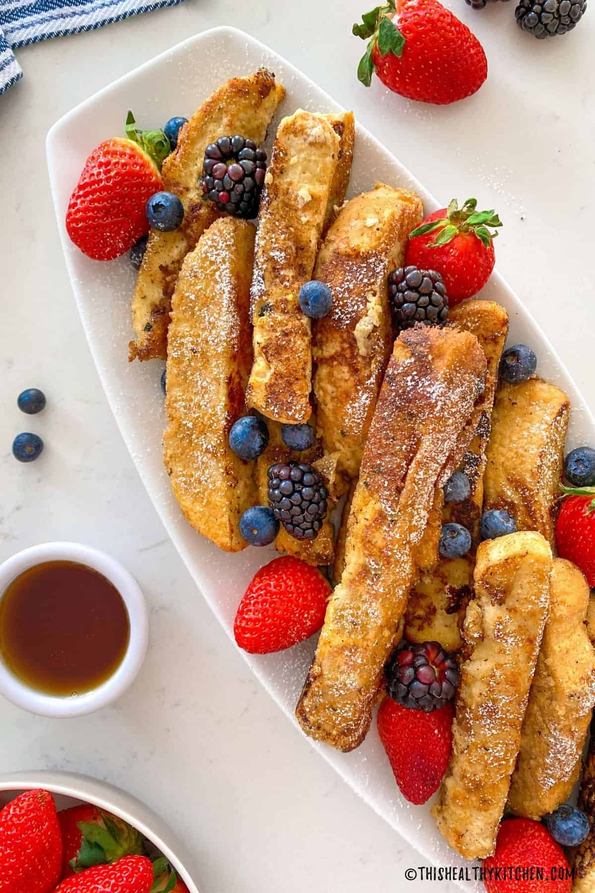 French toast sticks in white platter with strawberries, blueberries and black berries on top.