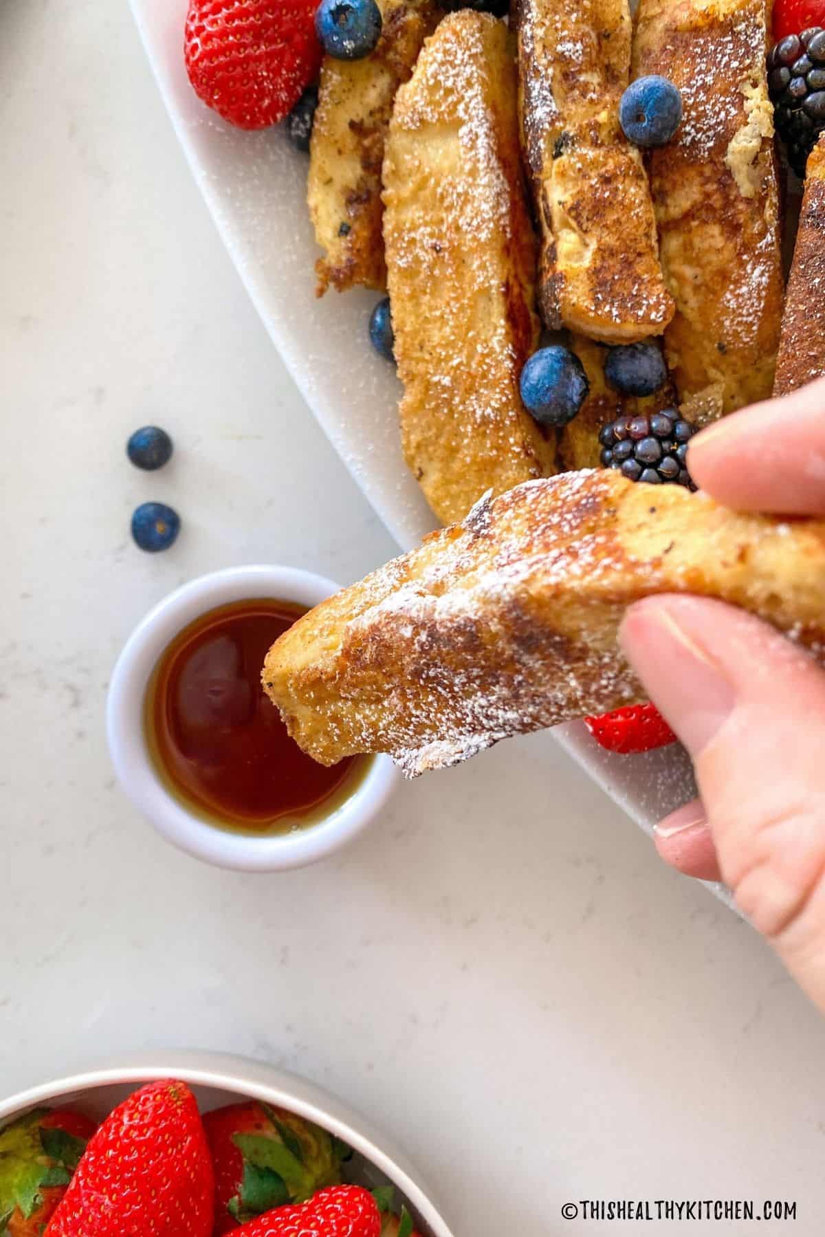 Hand dipping french toast stick into bowl of maple syrup.
