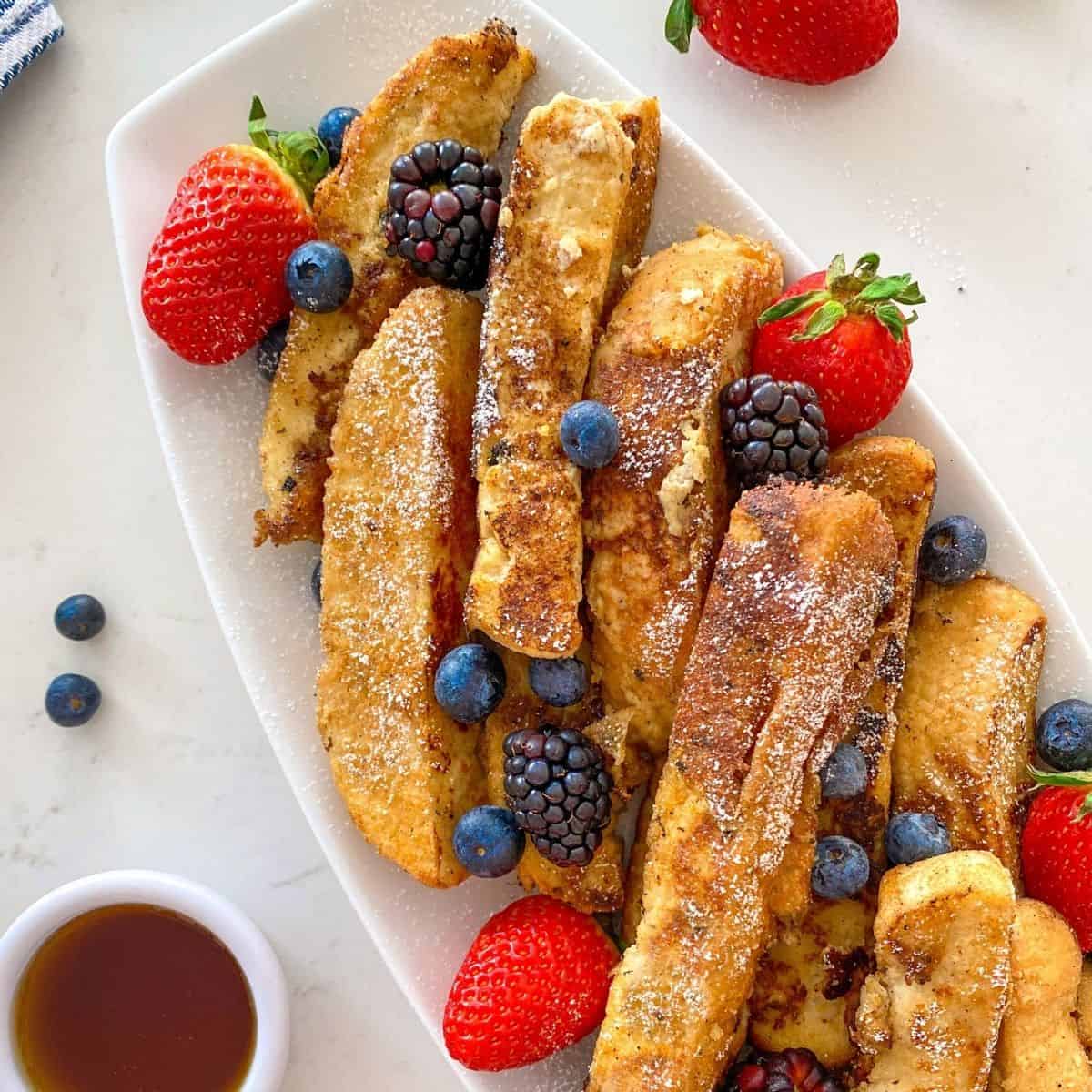French toast sticks in white platter with strawberries, blueberries and black berries on top.
