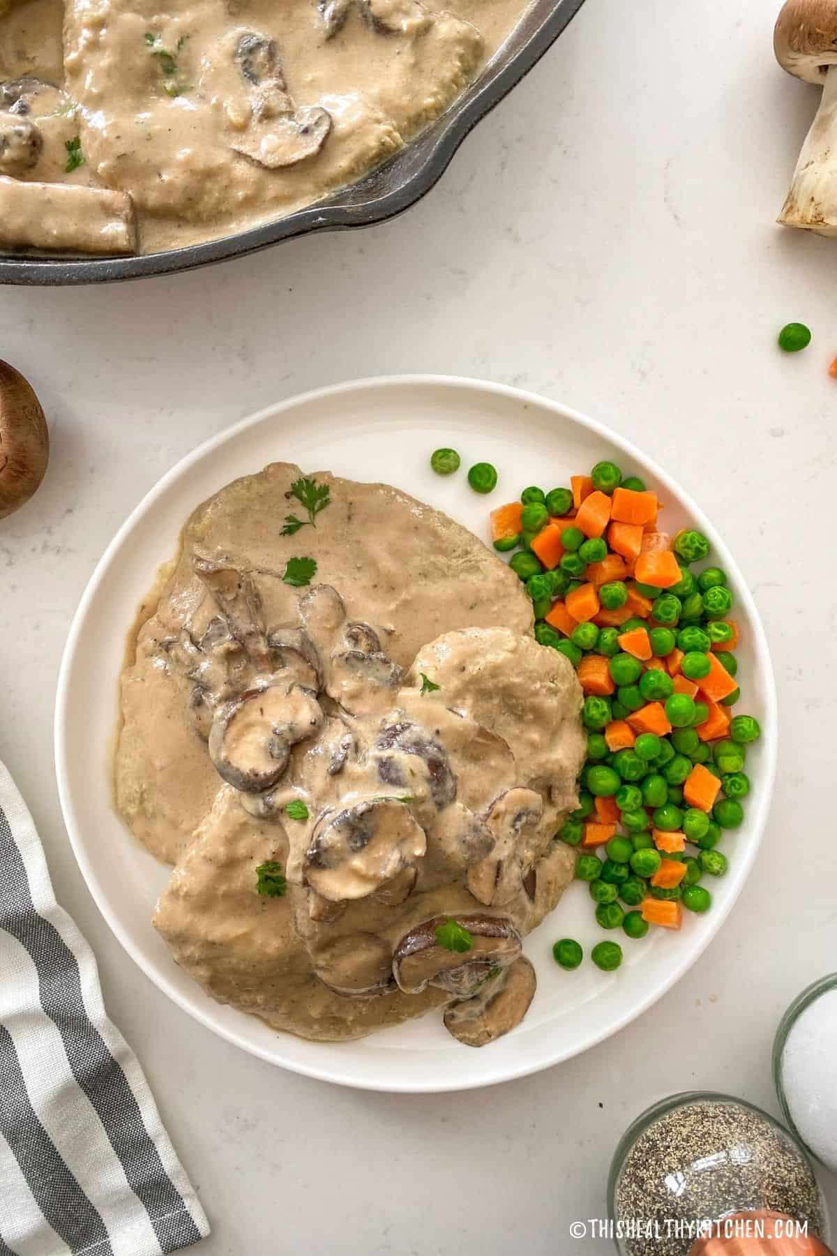 Vegan chicken cutlets on plate with mushroom sauce and peas and carrots on the side.