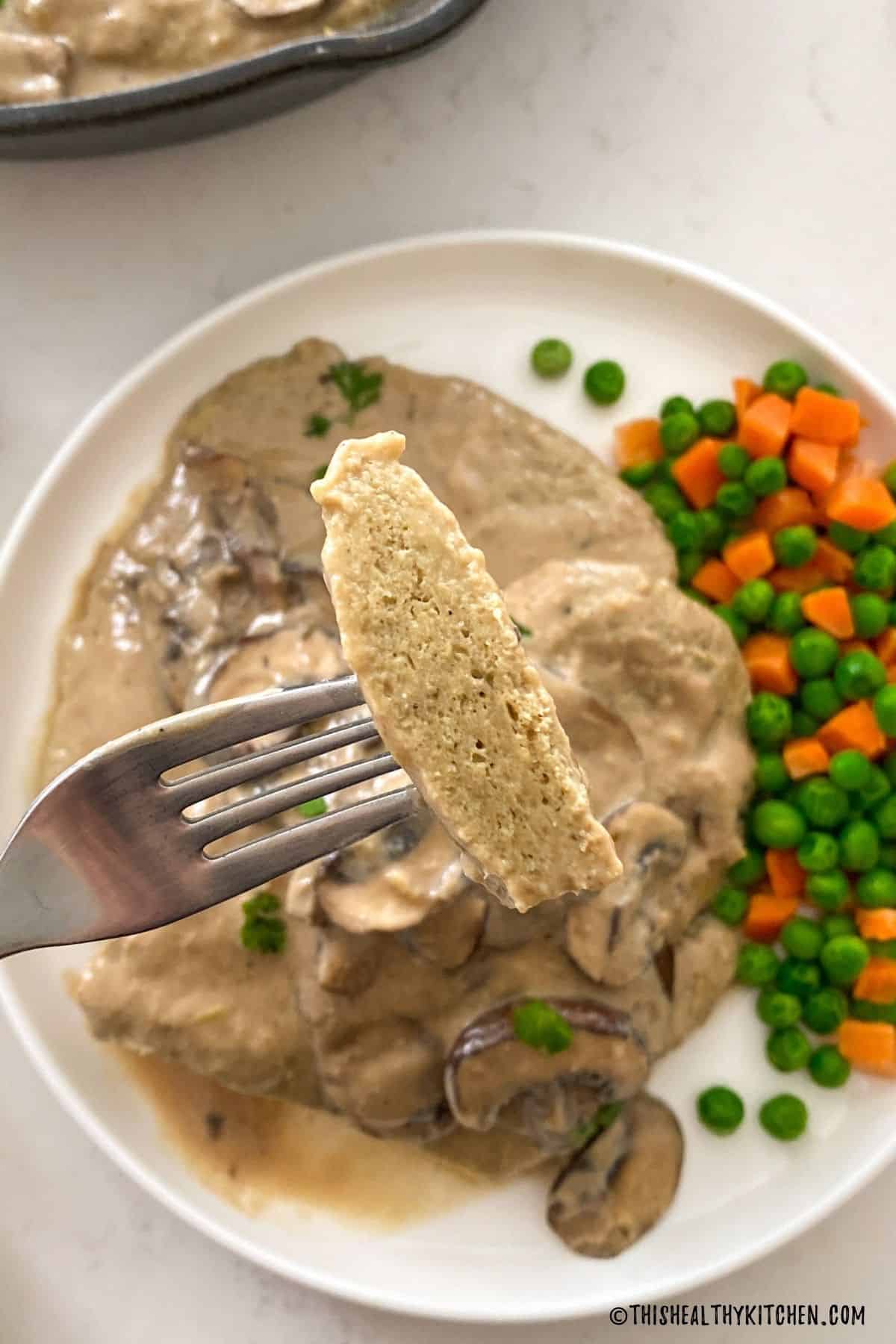 Fork holding up one bite of vegan chicken with plate of chicken and veggies below.
