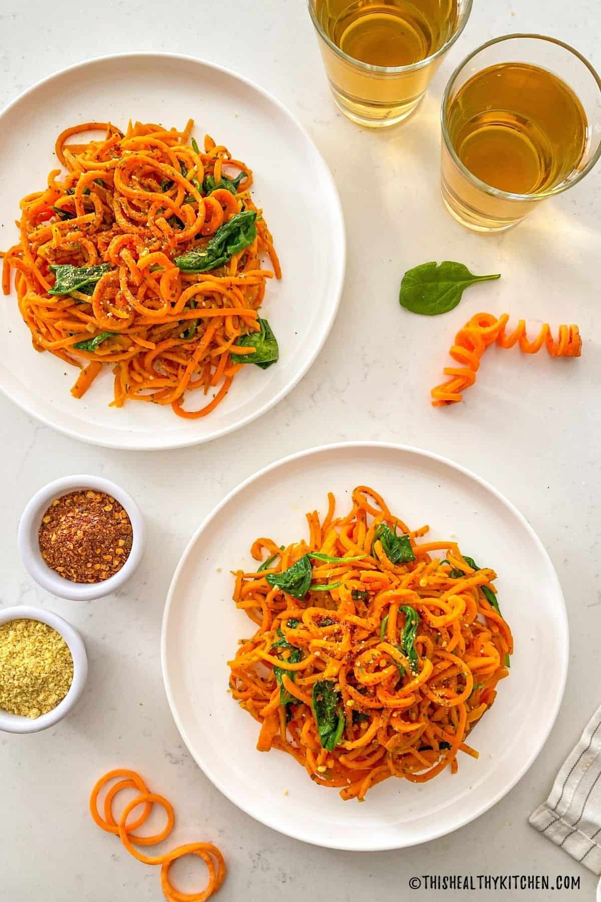 Two plates of sweet potato spaghetti with two glasses of wine and small bowl of chili flakes.