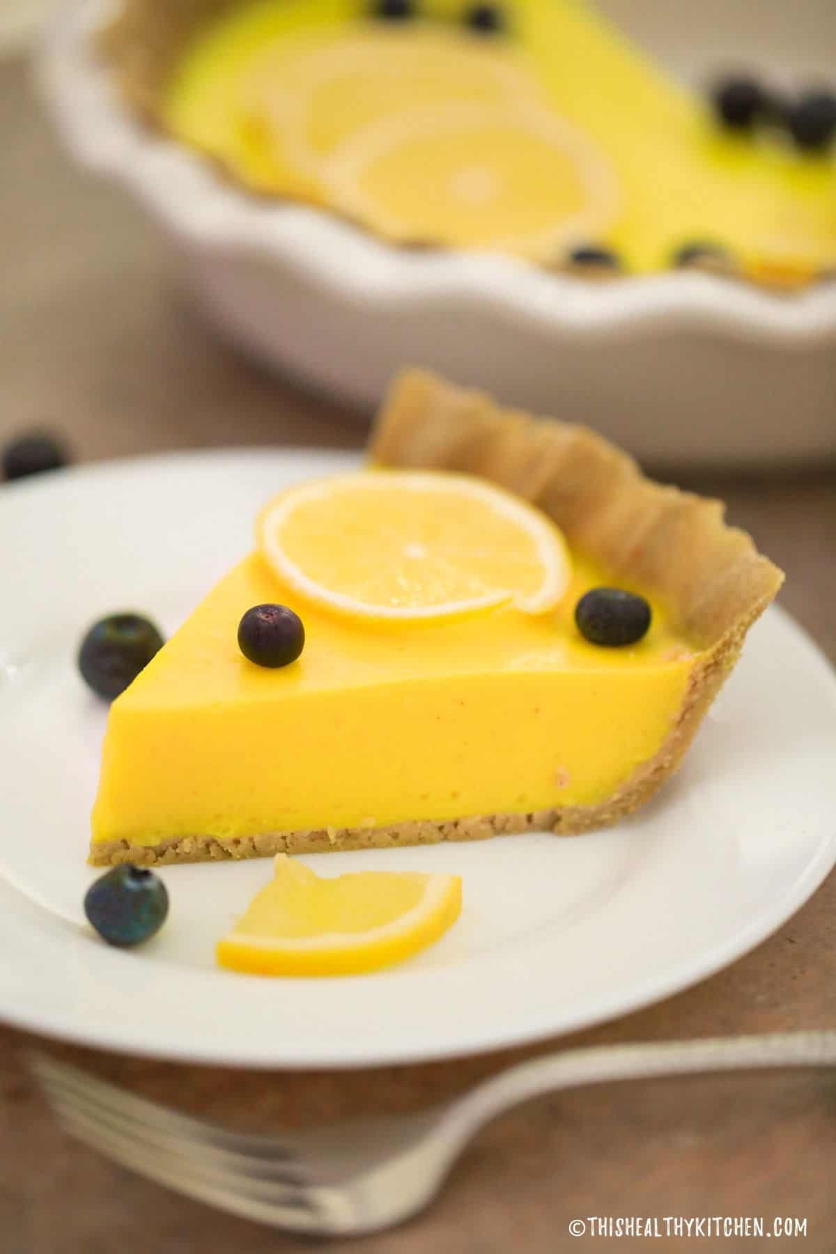 Slice of lemon pie in plate with remaining pie in the background.