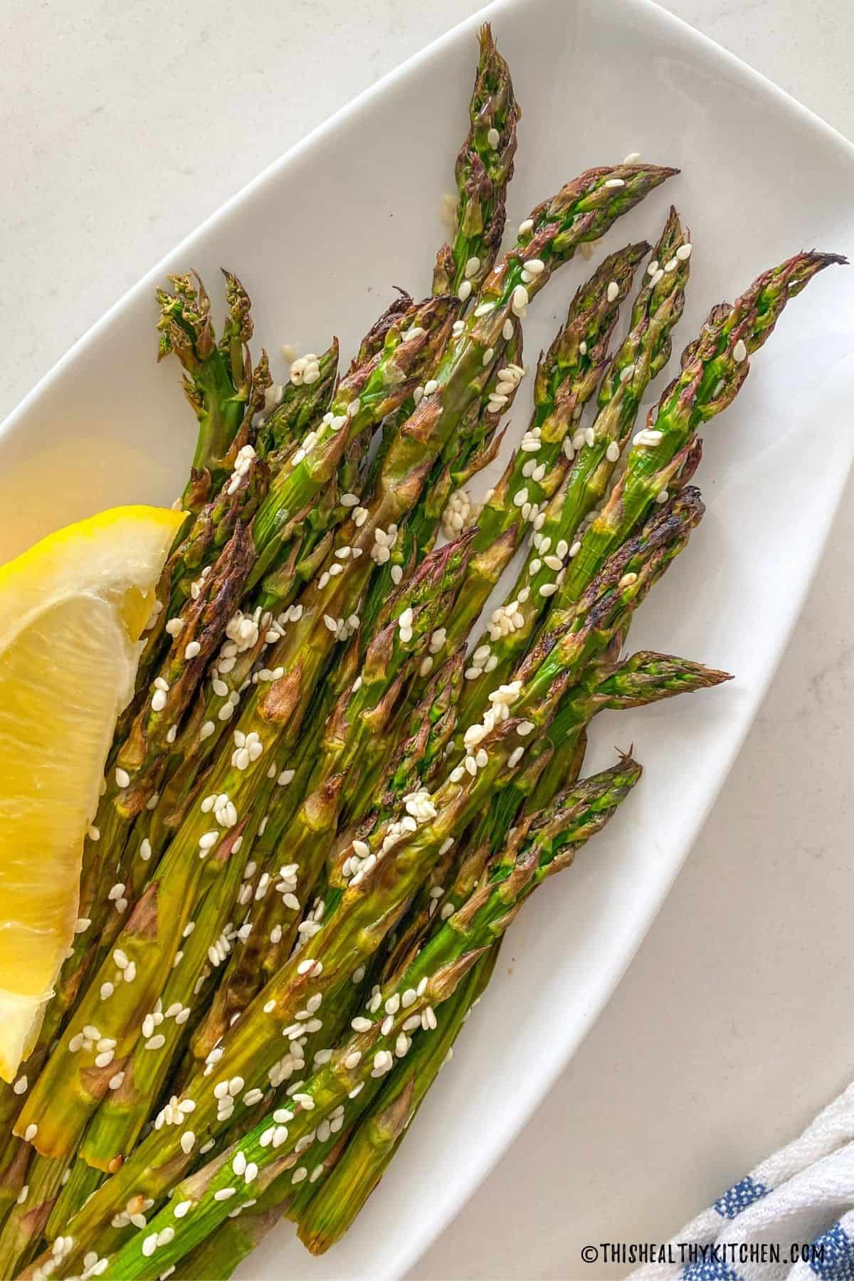 Asparagus in white serving dish with sesame seeds and lemon wedge.