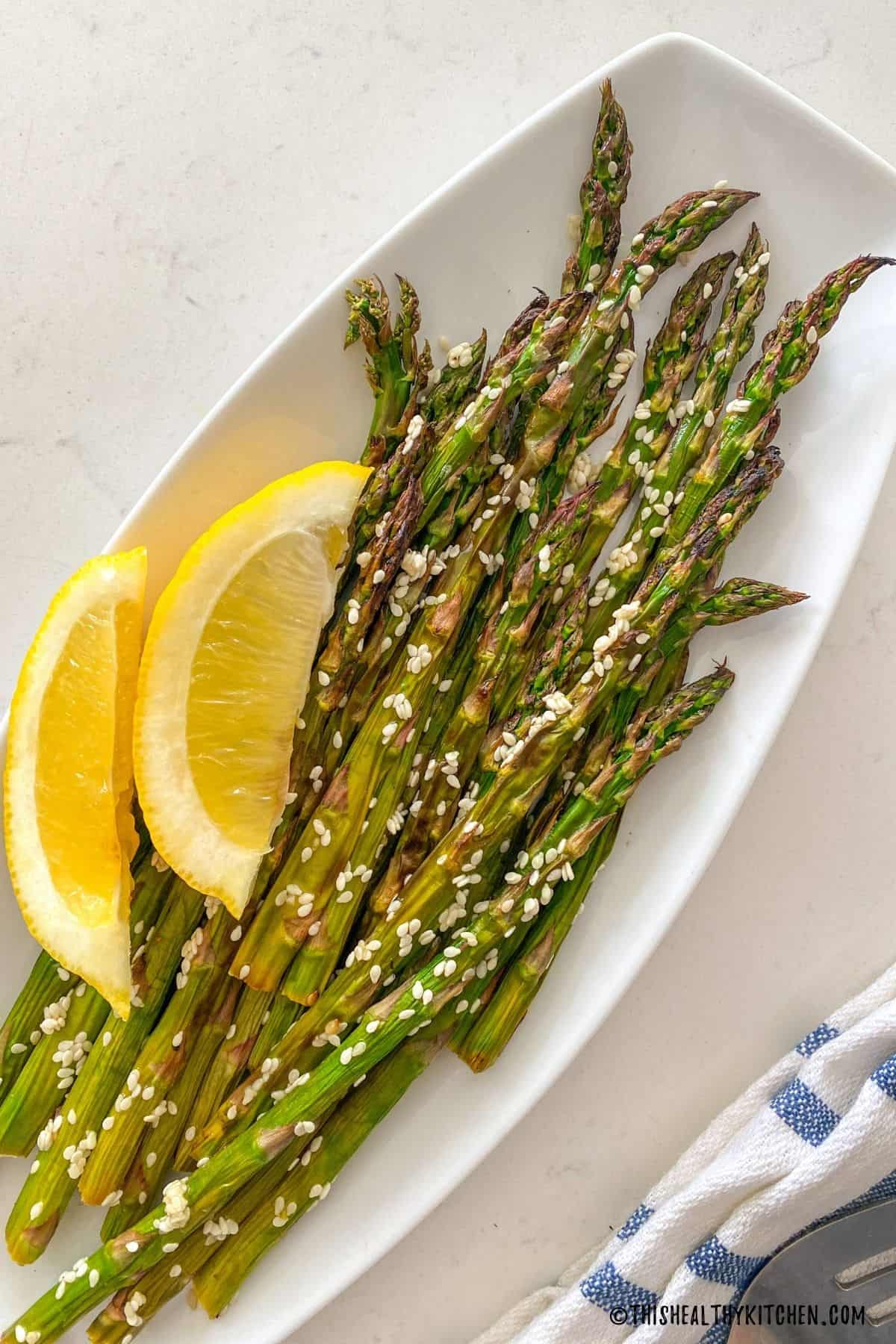 Roasted asparagus with sesame seeds and lemon wedges in white dish.