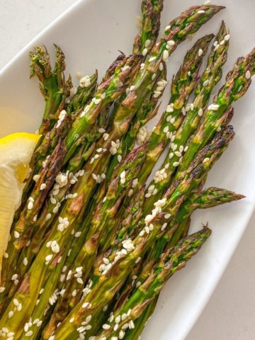 Asparagus in white serving dish with sesame seeds and lemon wedge.