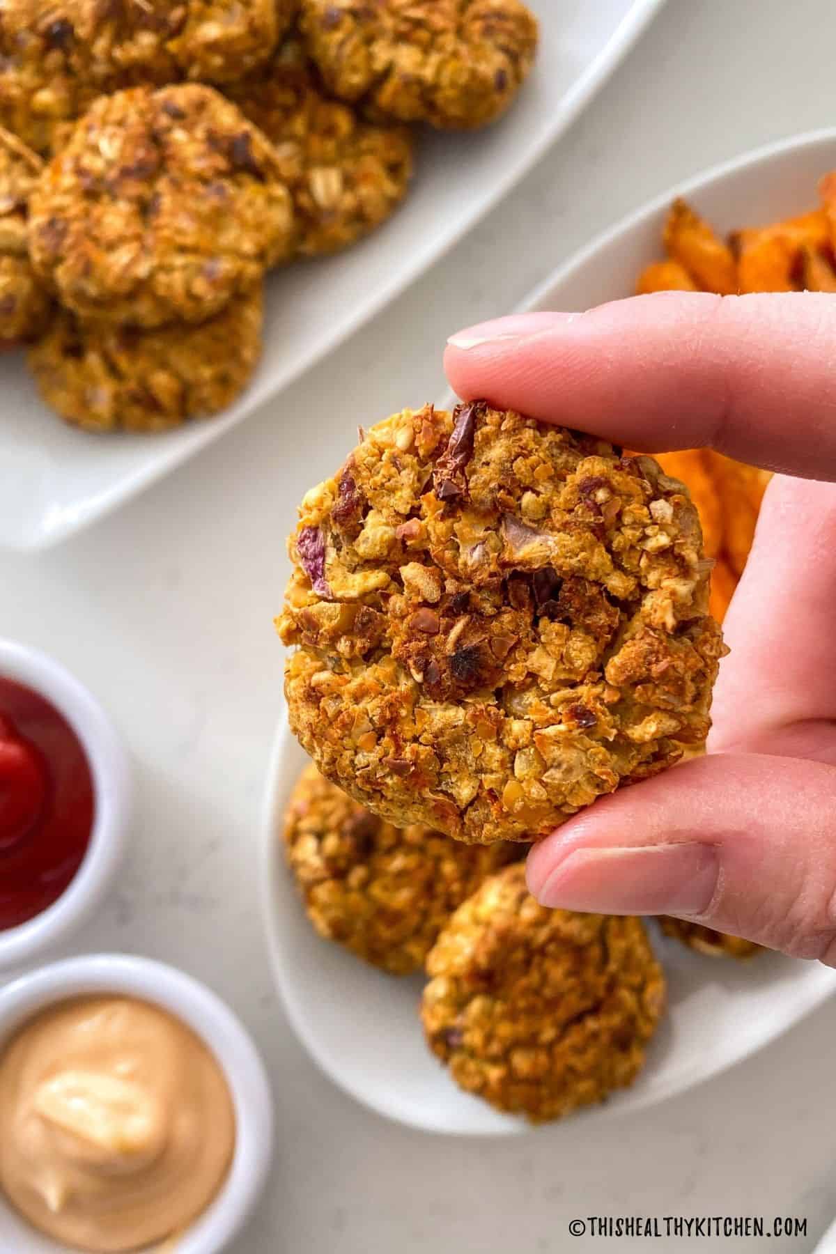 Hand holding up one vegan nugget above platter with more of them.
