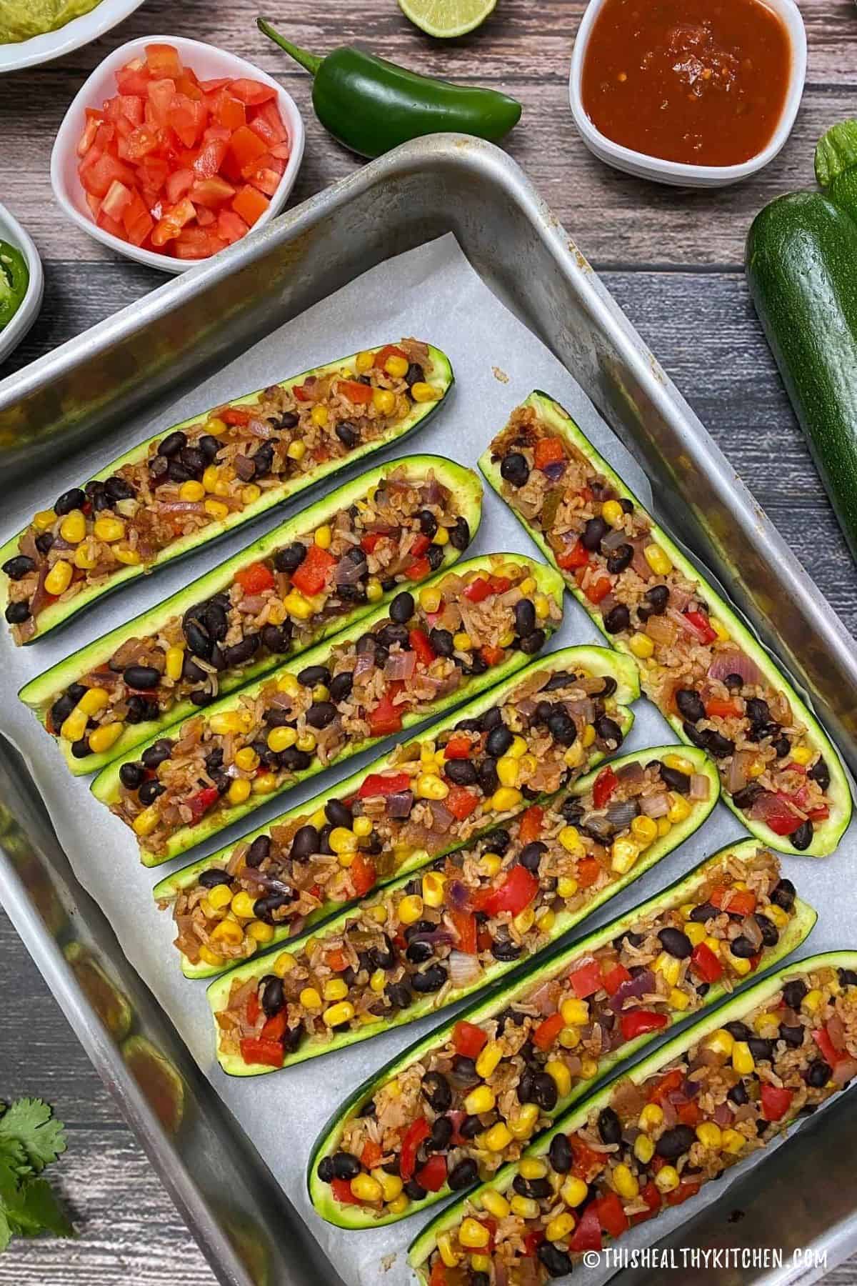Zucchini boats in large baking tray with beans, rice, peppers and corn inside.
