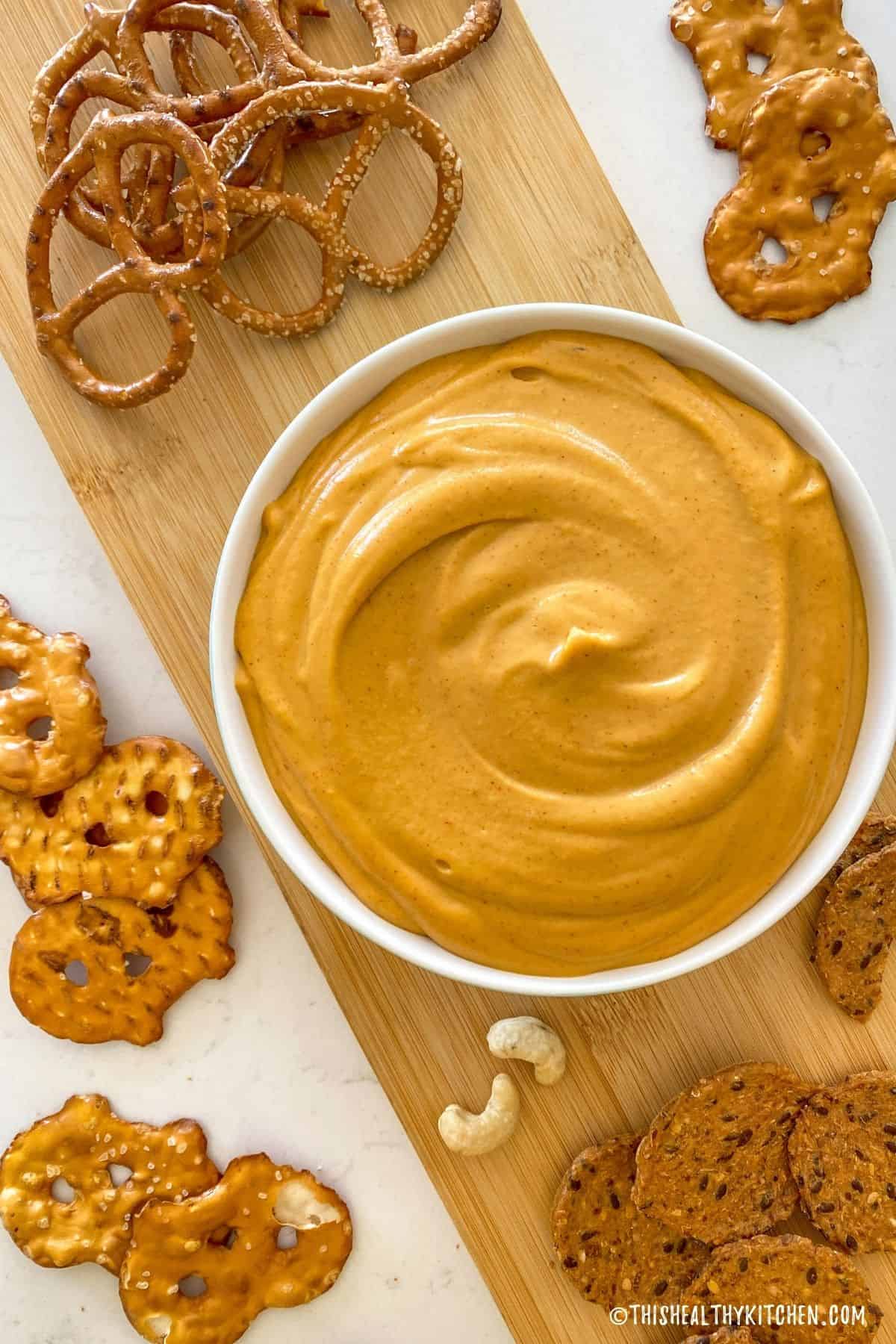 White bowl with orange dip inside and crackers and pretzels scattered around it.