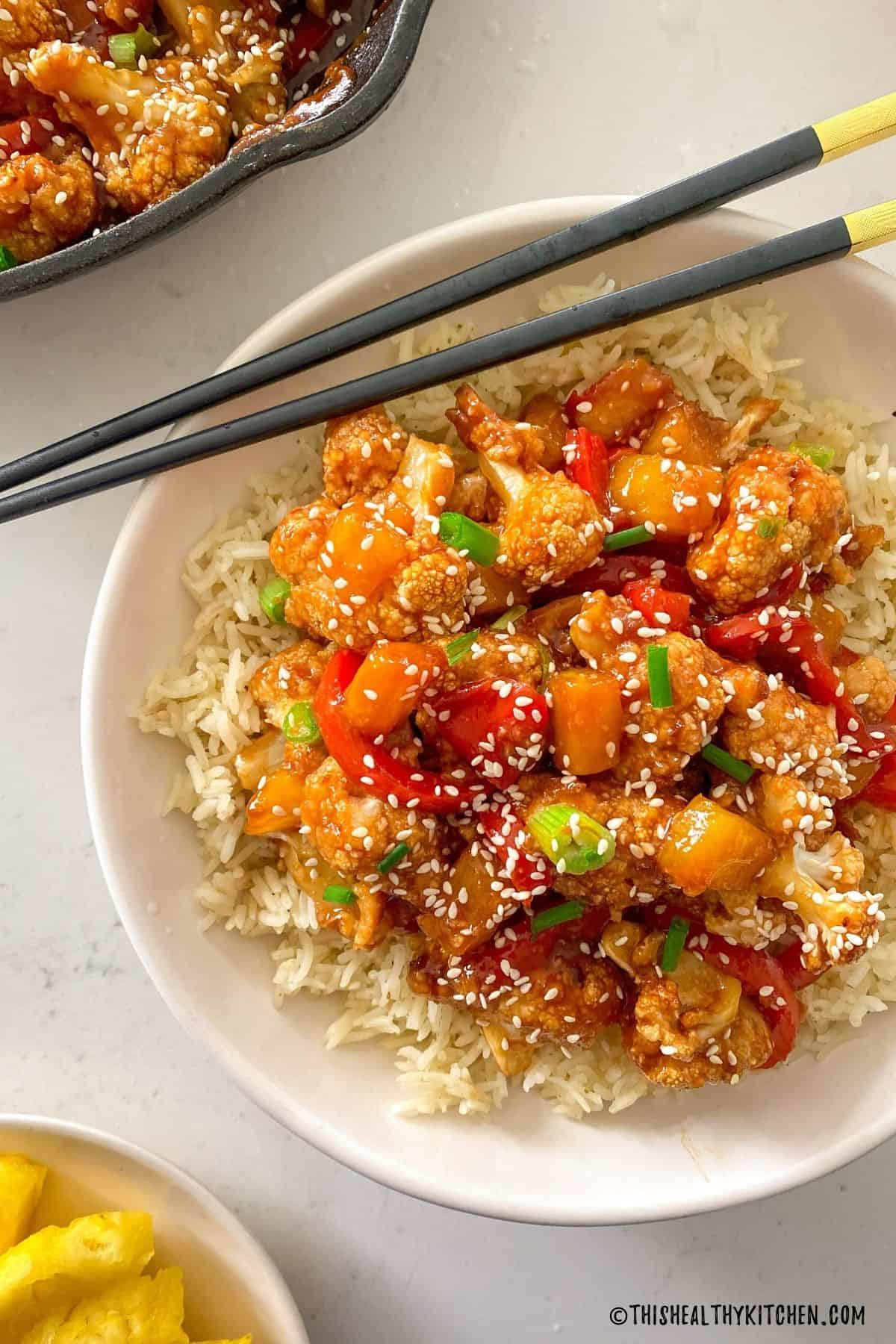 Sweet and sour cauliflower on top of rice in white bowl with black chopsticks on top.