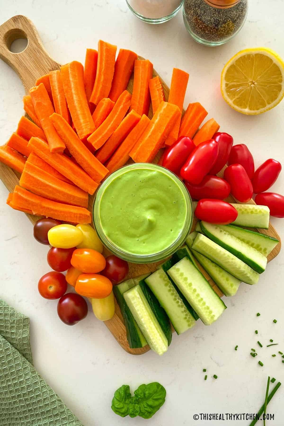 Cutting board with cucumber sticks, carrot sticks and cherry tomatoes around jar of green dip.