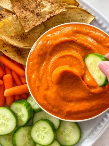 Bowl of red pepper hummus with cucumber, carrots and chips around it.