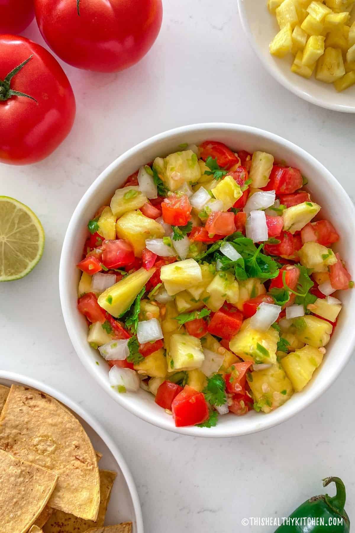 Pineapple pico de gallo in white bowl with plate of tortilla chips beside it.