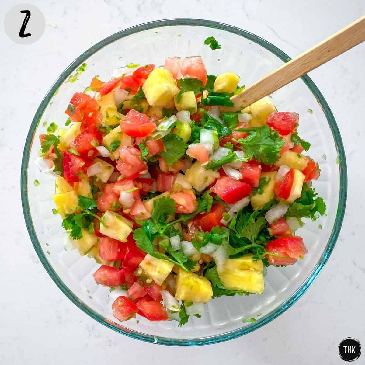 Glass mixing bowl filled with diced pineapple, tomato, jalapeno, cilantro and white onion.