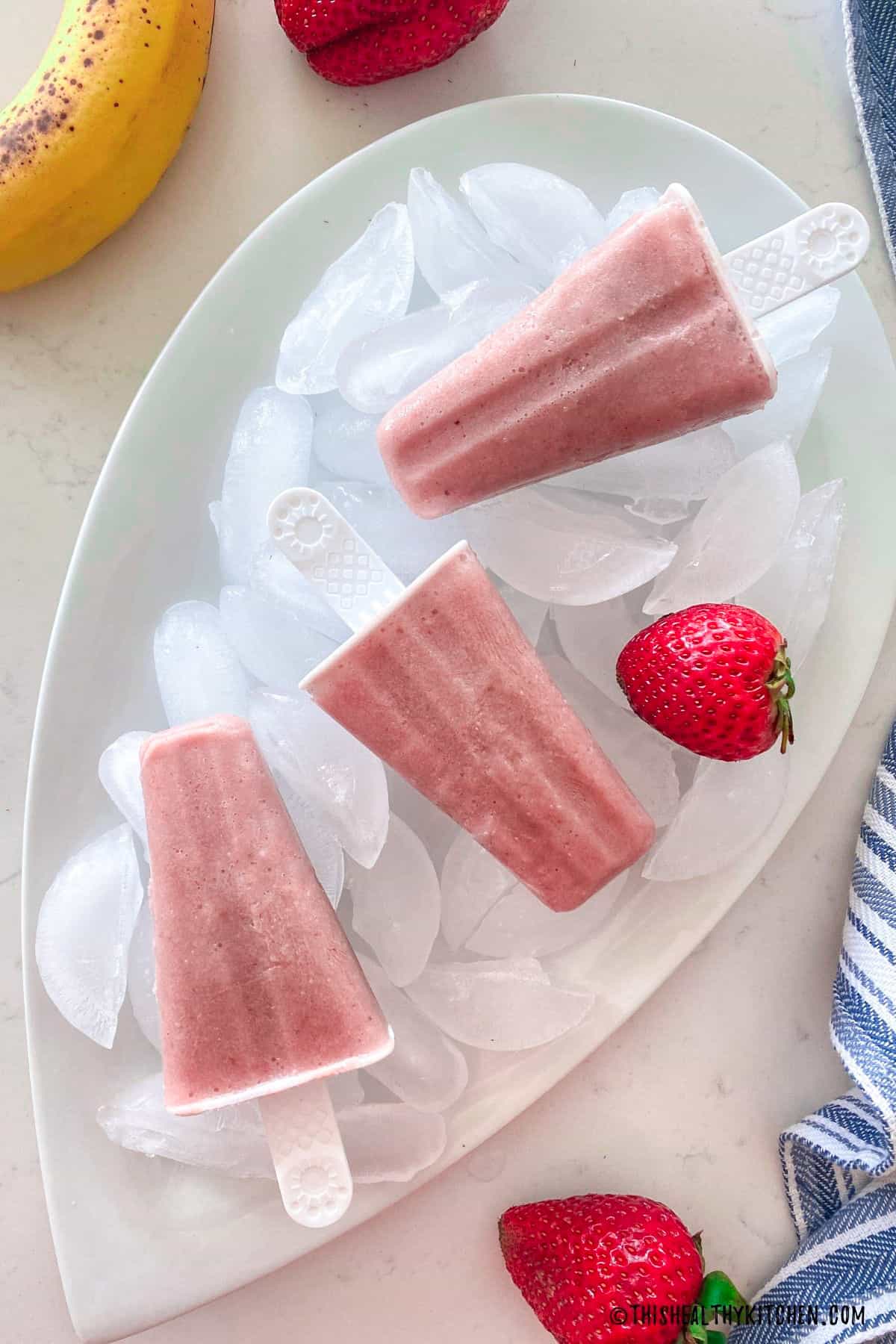 Three pink popsicles resting on ice cubes with strawberries scattered around.