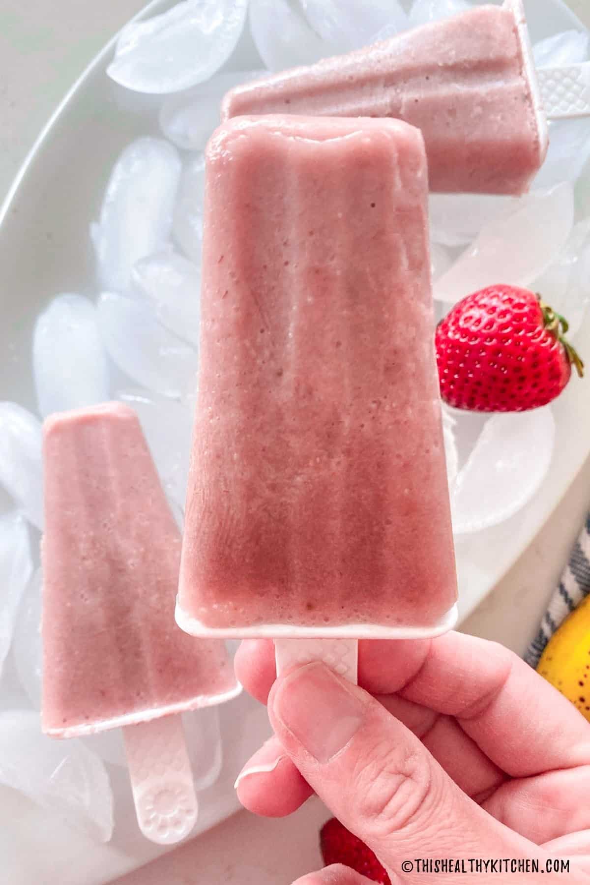 Hand holding up pink popsicle with more on tray below.