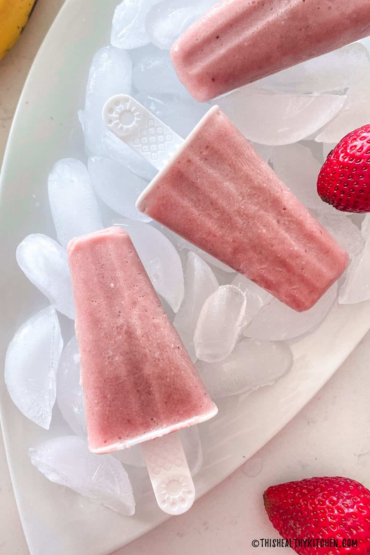 Pink popsicles on tray of ice cubes with fresh strawberry on the side.
