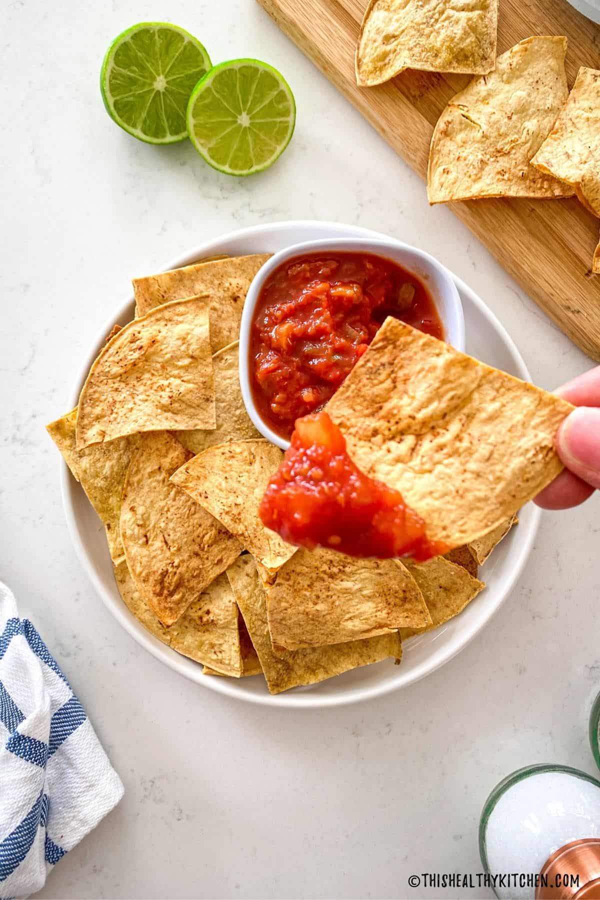 Chip dunked with red salsa being held above plate with more chips below.