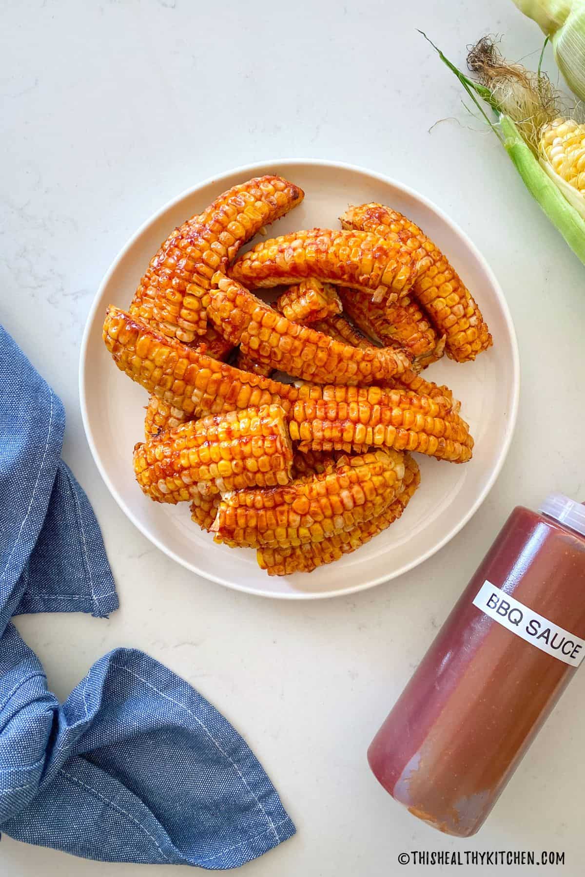 BBQ sauce covered corn on white plate with bottle of bbq sauce beside it.